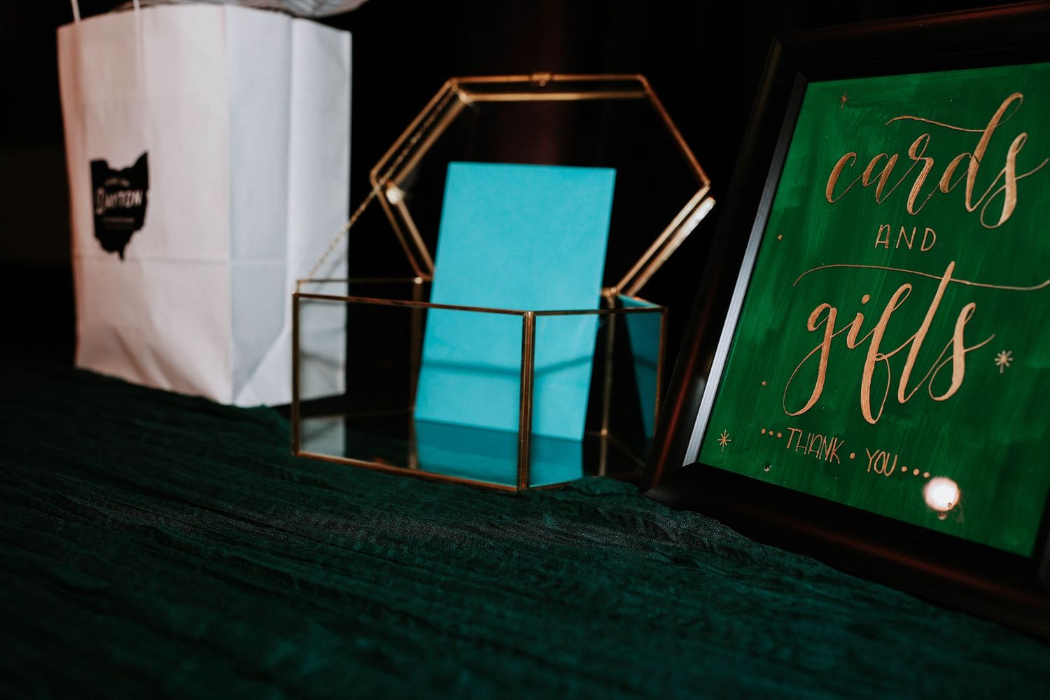 Elegant wedding cards and gifts table with gold geometric box, teal accents, and green velvet backdrop.