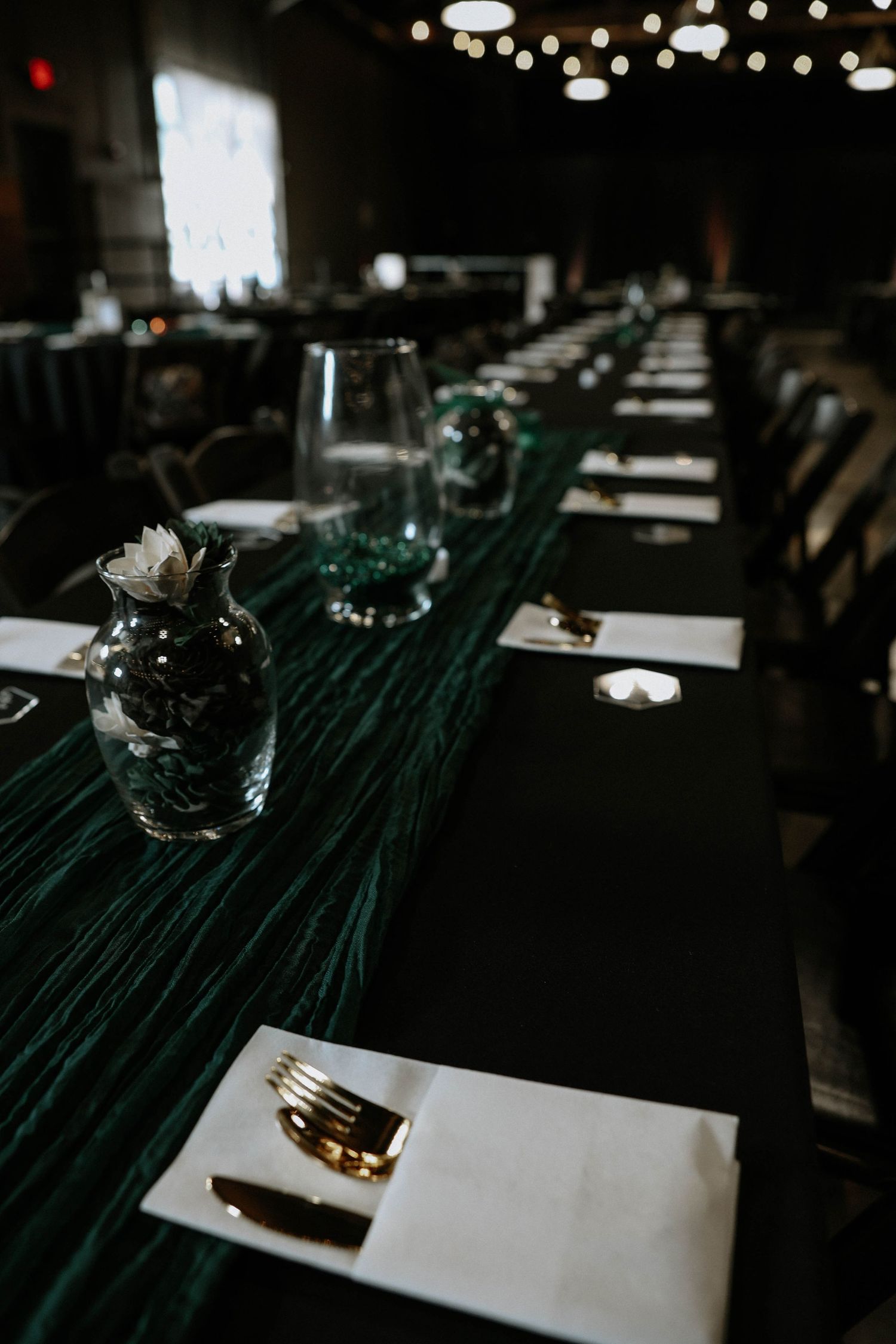 Elegant dark reception hall with long green velvet table runner, gold cutlery, crystal vases, and string lights above.