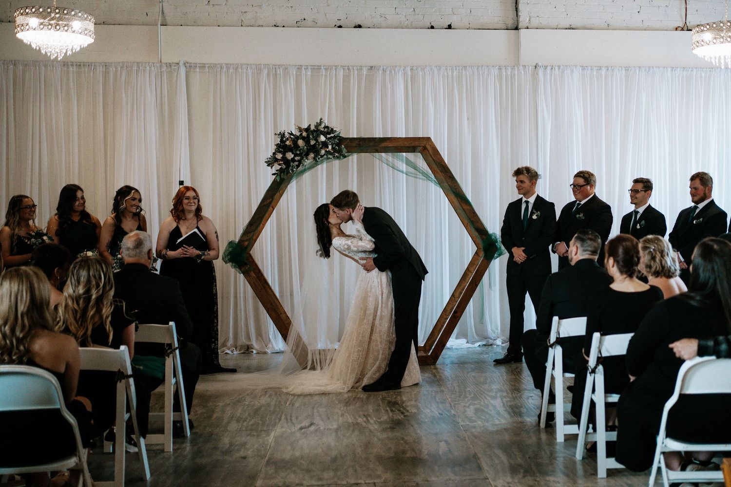 Bride and groom share first kiss under hexagonal wooden arch at elegant indoor wedding ceremony.