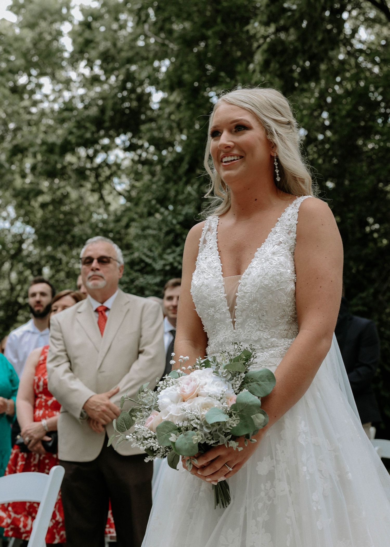 Radiant bride in a beaded V-neck wedding gown holding a bouquet of peonies and eucalyptus at outdoor ceremony.