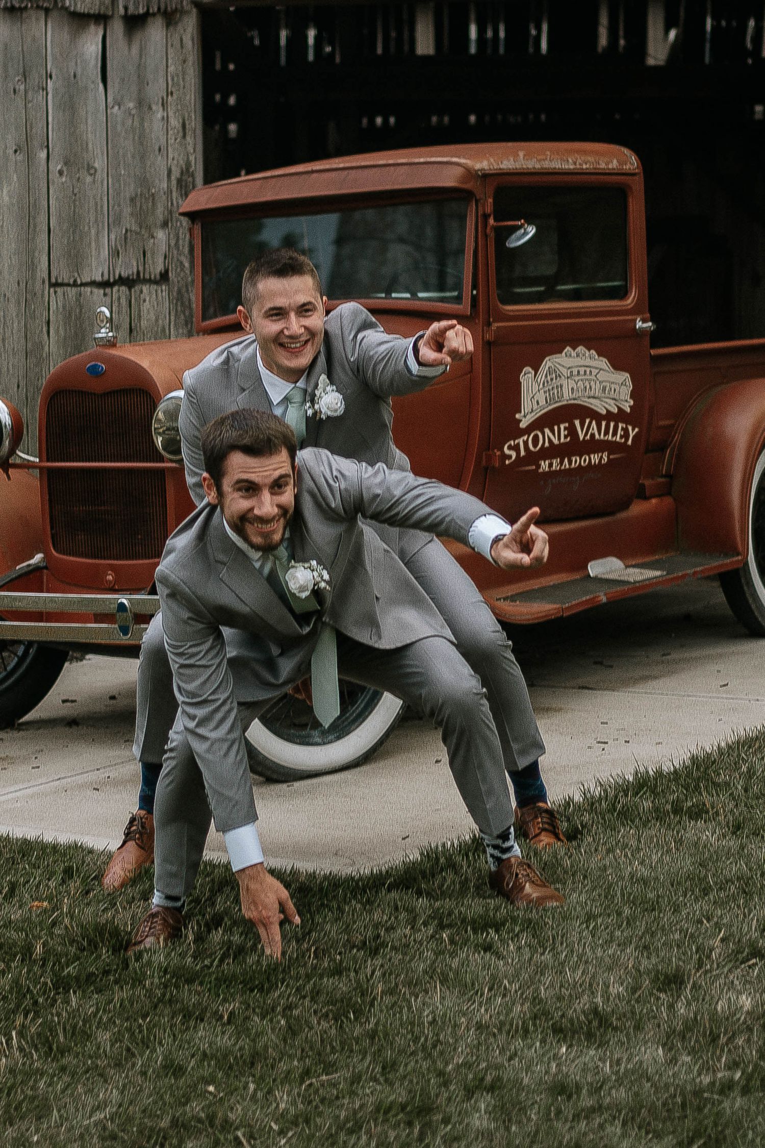 Two groomsmen in gray suits posing playfully in front of a vintage Stone Valley Meadows red truck.