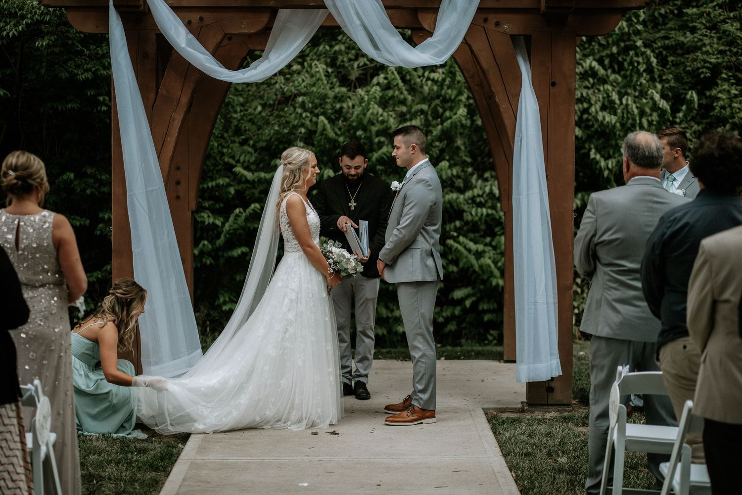 Bride and groom exchange vows under a wooden arch with white draping at an outdoor wedding ceremony.