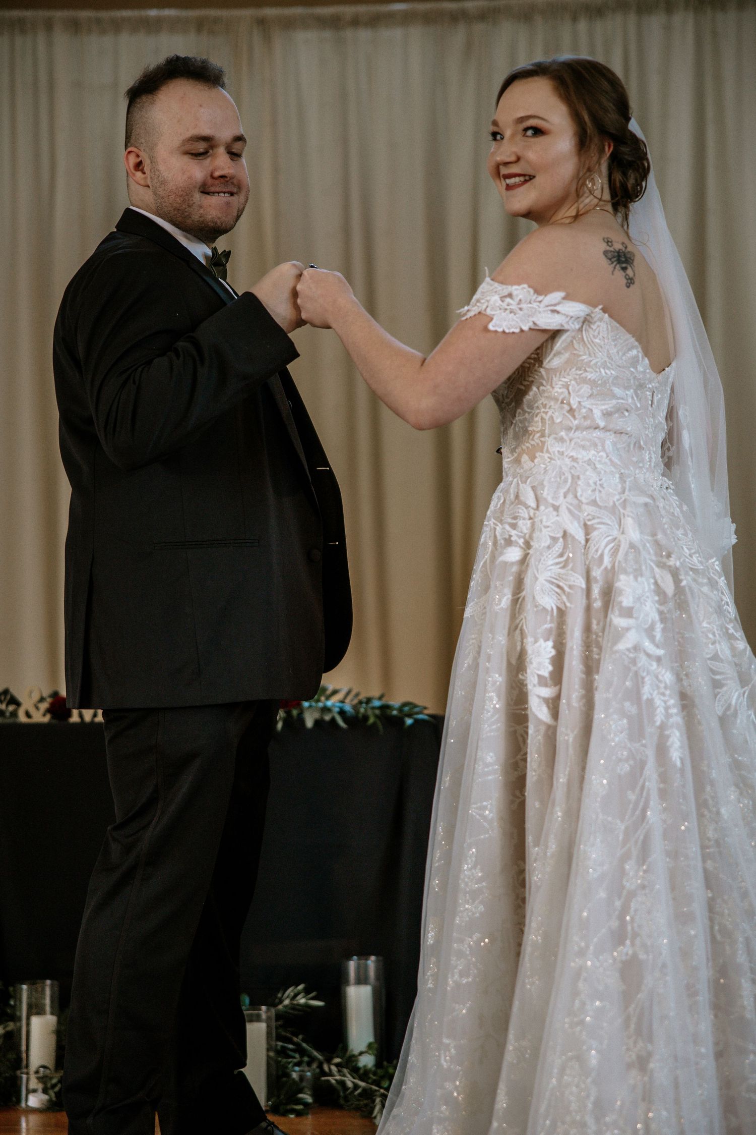 Smiling bride in lace off-shoulder gown fist bumps groom in black tuxedo at wedding altar at Urban Artifact in Cincinnati, Ohio