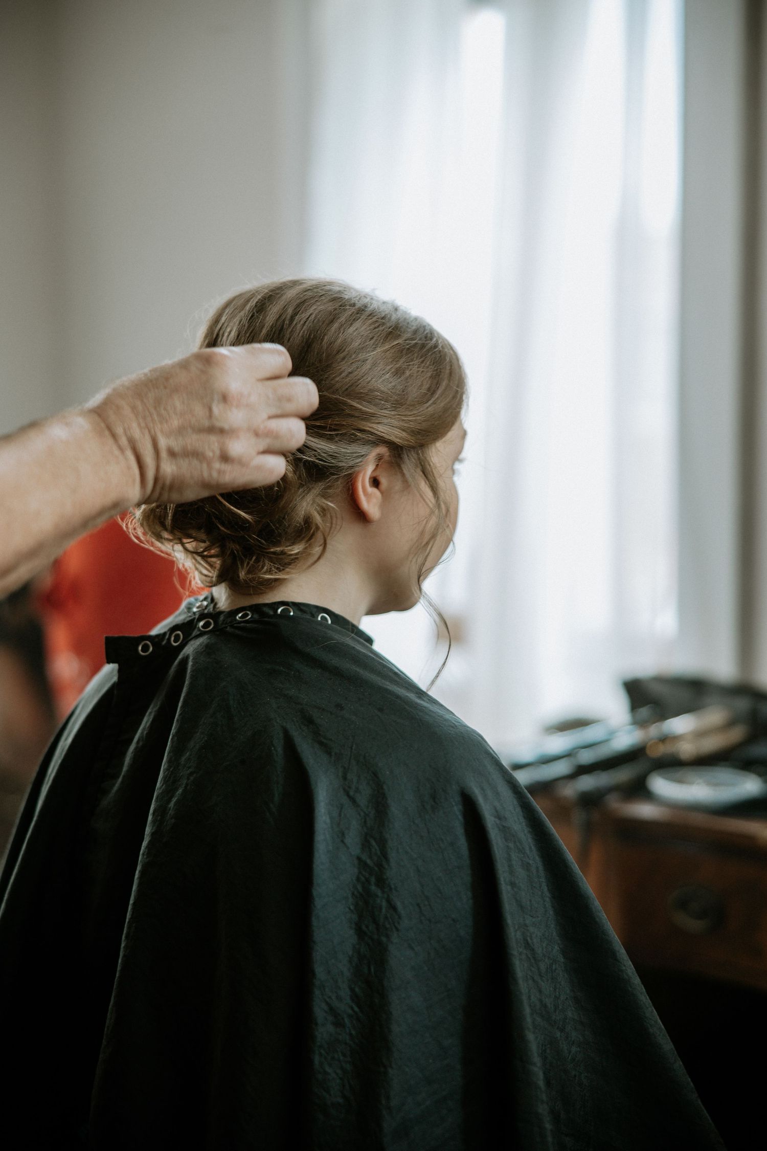 Hair stylist styling bride's updo hairstyle while she sits in a black cape near a window at Urban Artifact in Cincinnati, Ohio