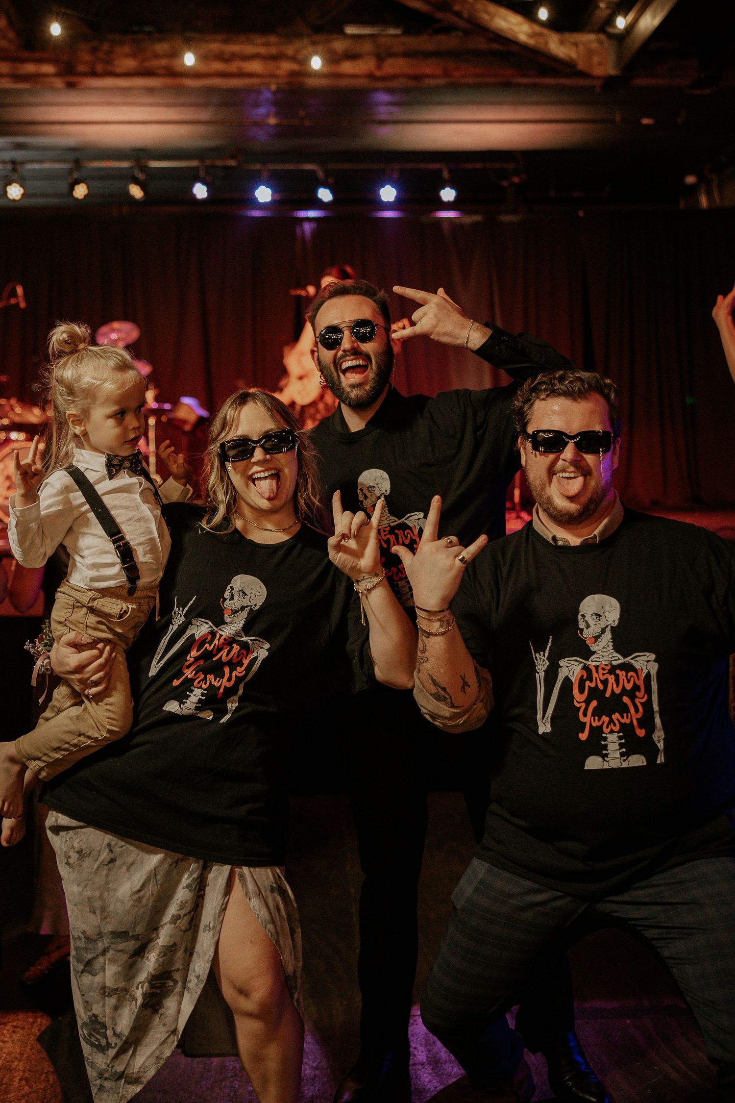 Wedding guests in matching skeleton T-shirts pose with rock hand signs at a reception.
