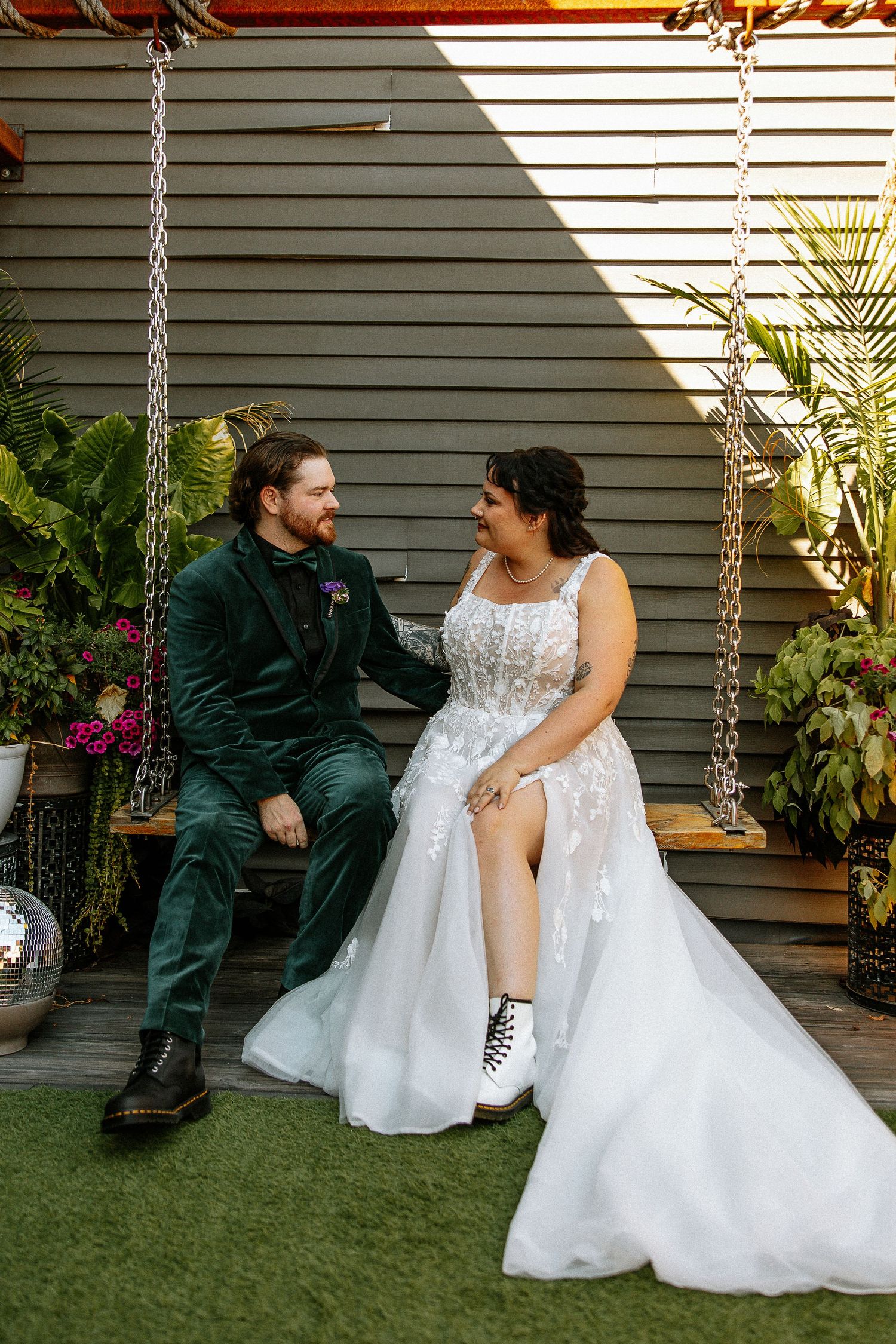 Bride in lace wedding gown with combat boots sits with groom in green velvet suit on garden swing.