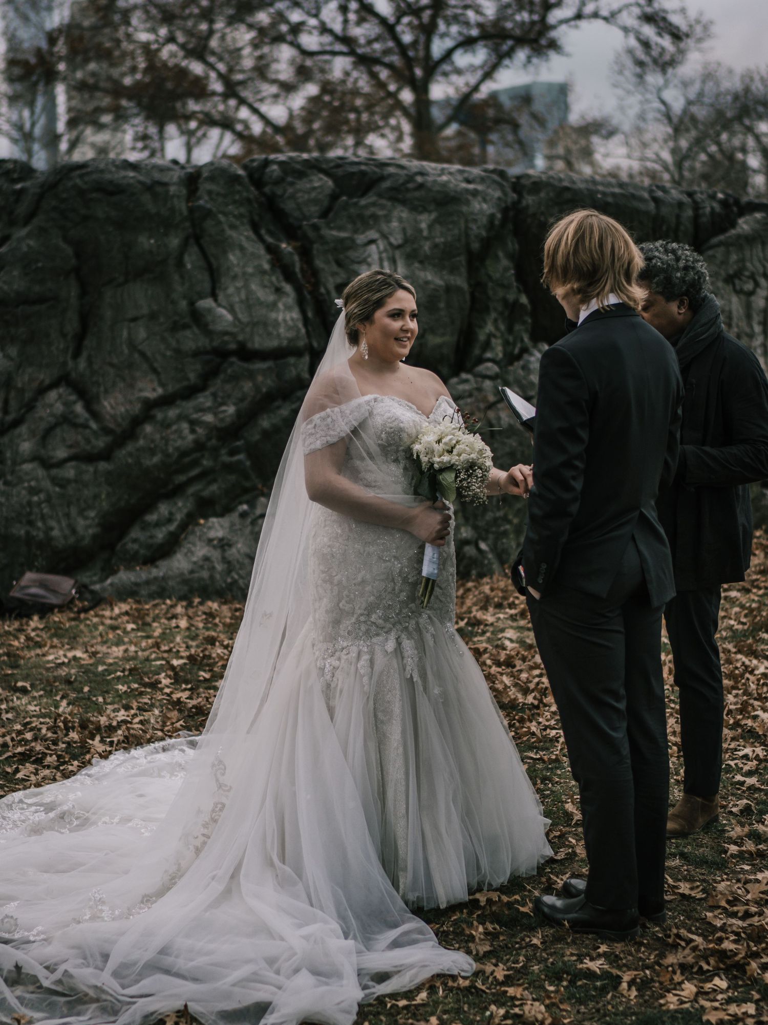 Smiling bride in lace mermaid gown reads vows to groom near rocky outcrop in Central Park autumn setting.
