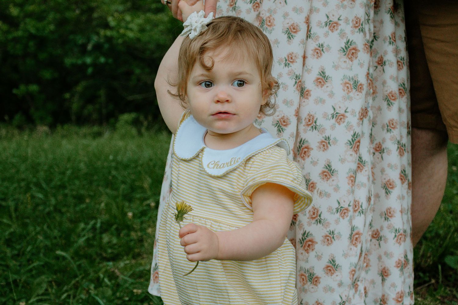 Sweet toddler girl in monogrammed yellow dress holding flower beside mother's floral skirt at French Park in Cincinnati, Ohio
