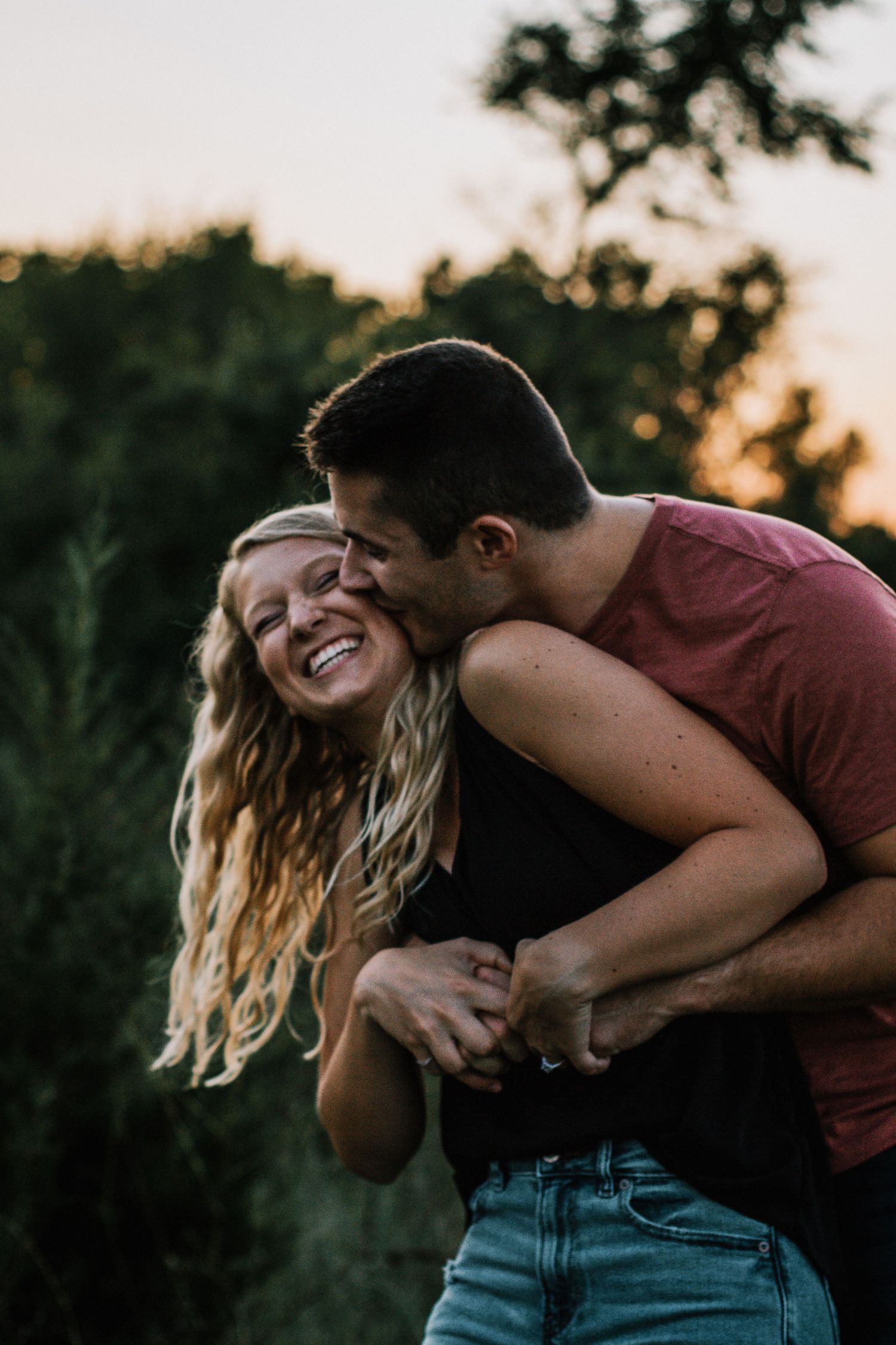 A joyful couple embraces outdoors at sunset, the man kissing the woman's cheek as she laughs happily in Cincinnati, Ohio