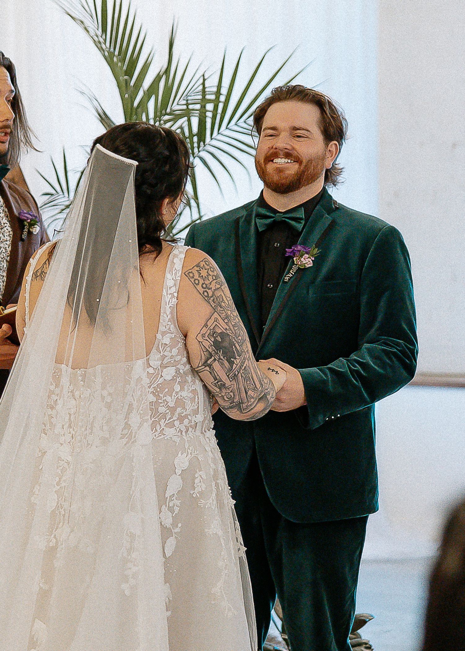Smiling groom in green velvet tuxedo holding bride's tattooed hands at wedding altar.