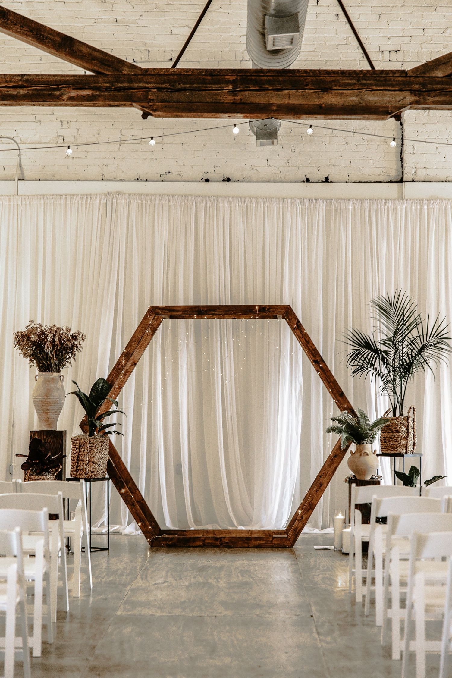 Hexagonal wooden wedding arch with tropical plants flanking white draped ceremony space with white chairs.