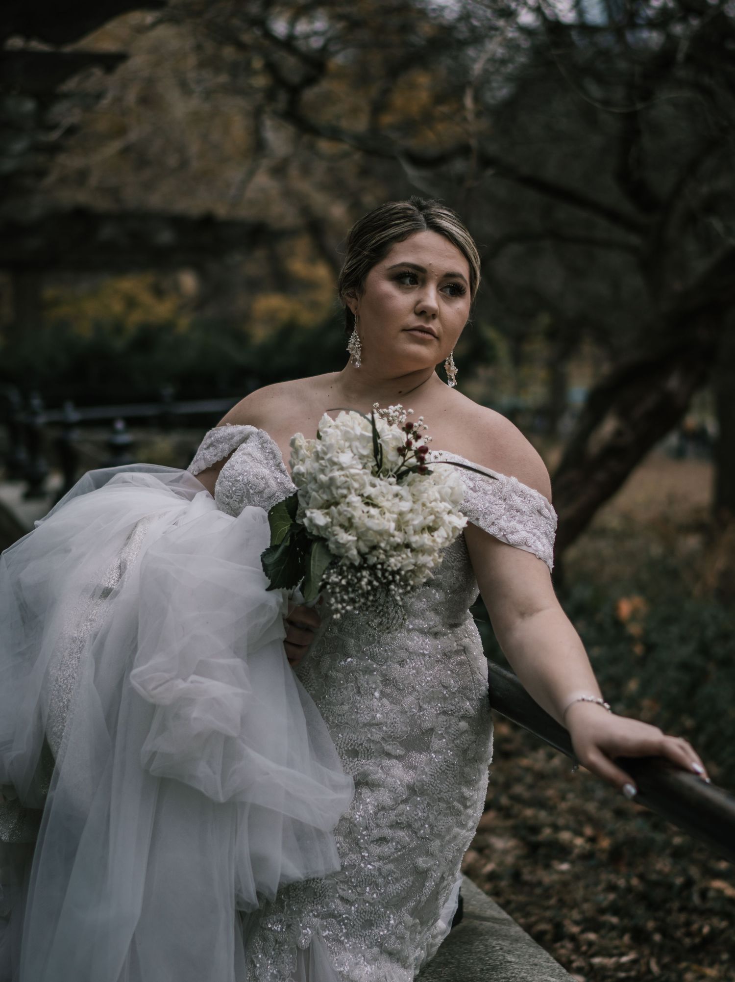 Elegant bride in off-shoulder lace mermaid gown holds white bouquet while posing near trees in Central Park.