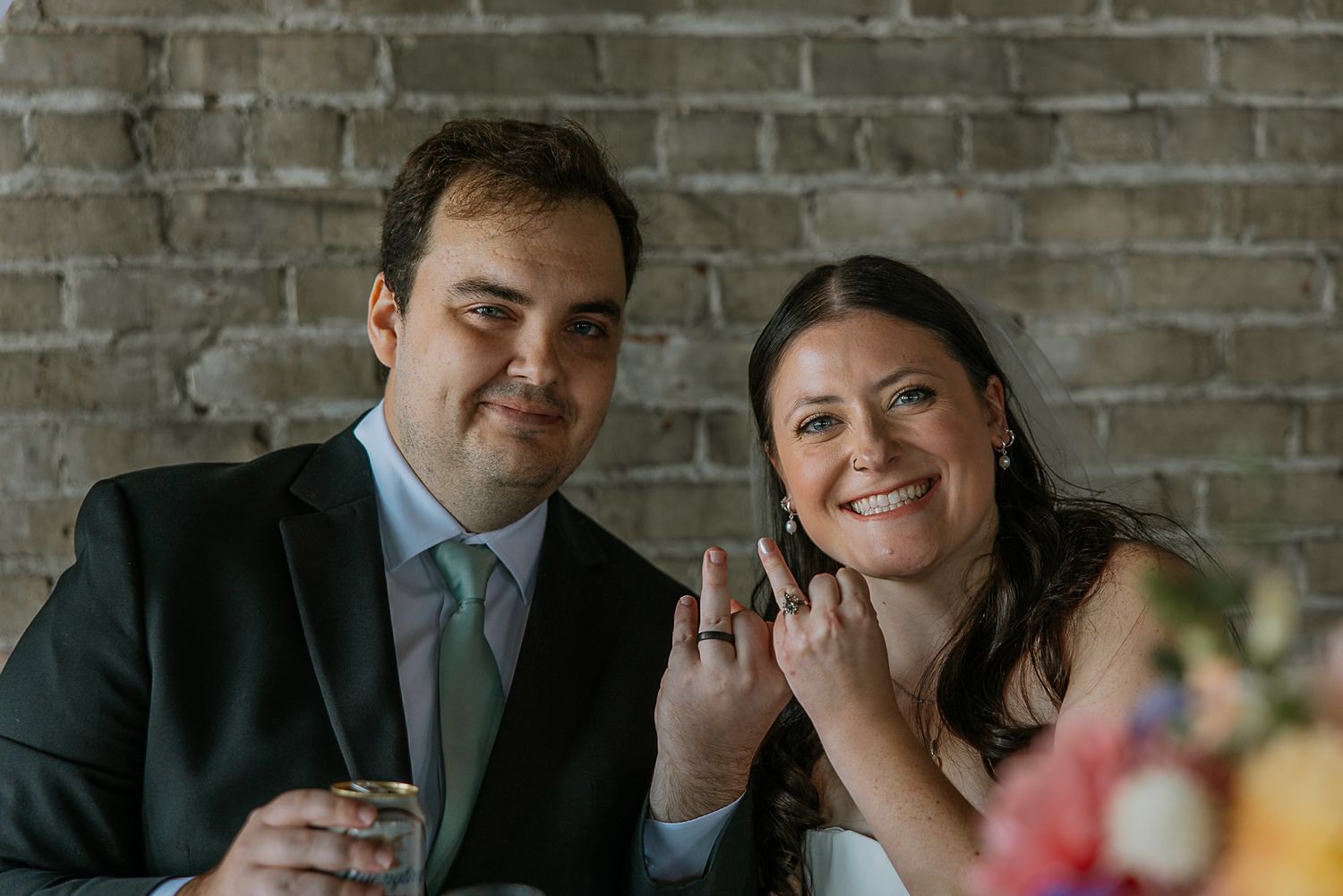 Newlyweds smile and show off wedding rings while seated at reception table against brick wall.