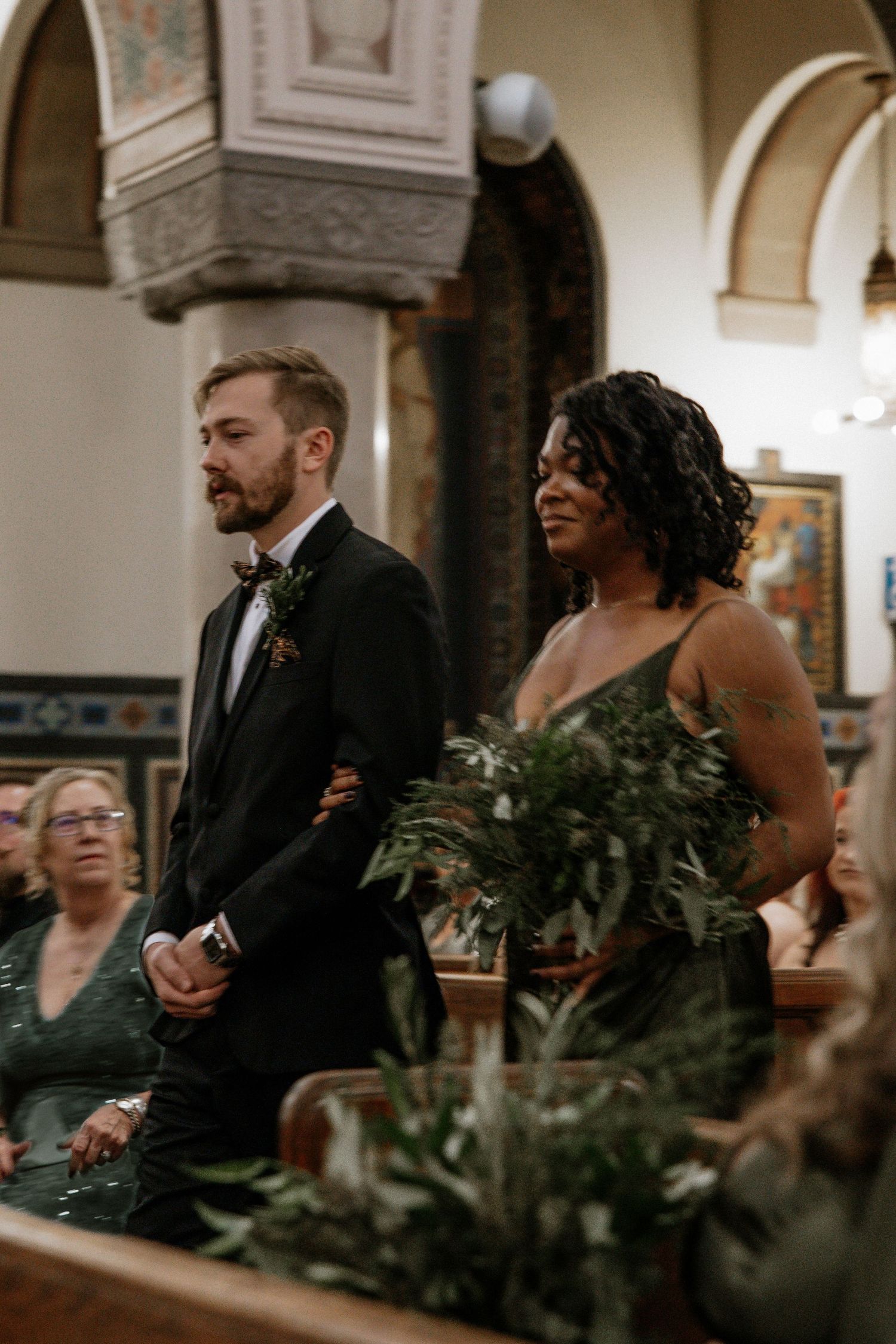 Groomsman in black tuxedo escorts bridesmaid in green dress down ornate church aisle at wedding in Cincinnati, Ohio