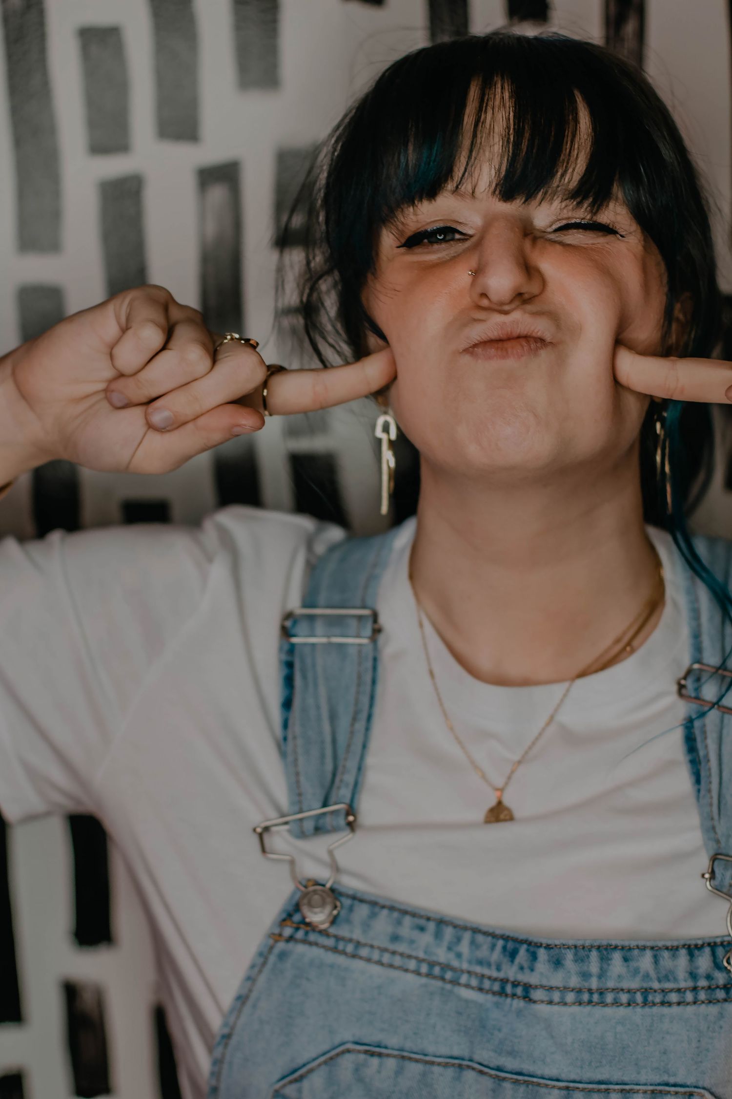 Young woman in denim overalls making a playful face, pressing her cheeks with fingers against a graphic wall.