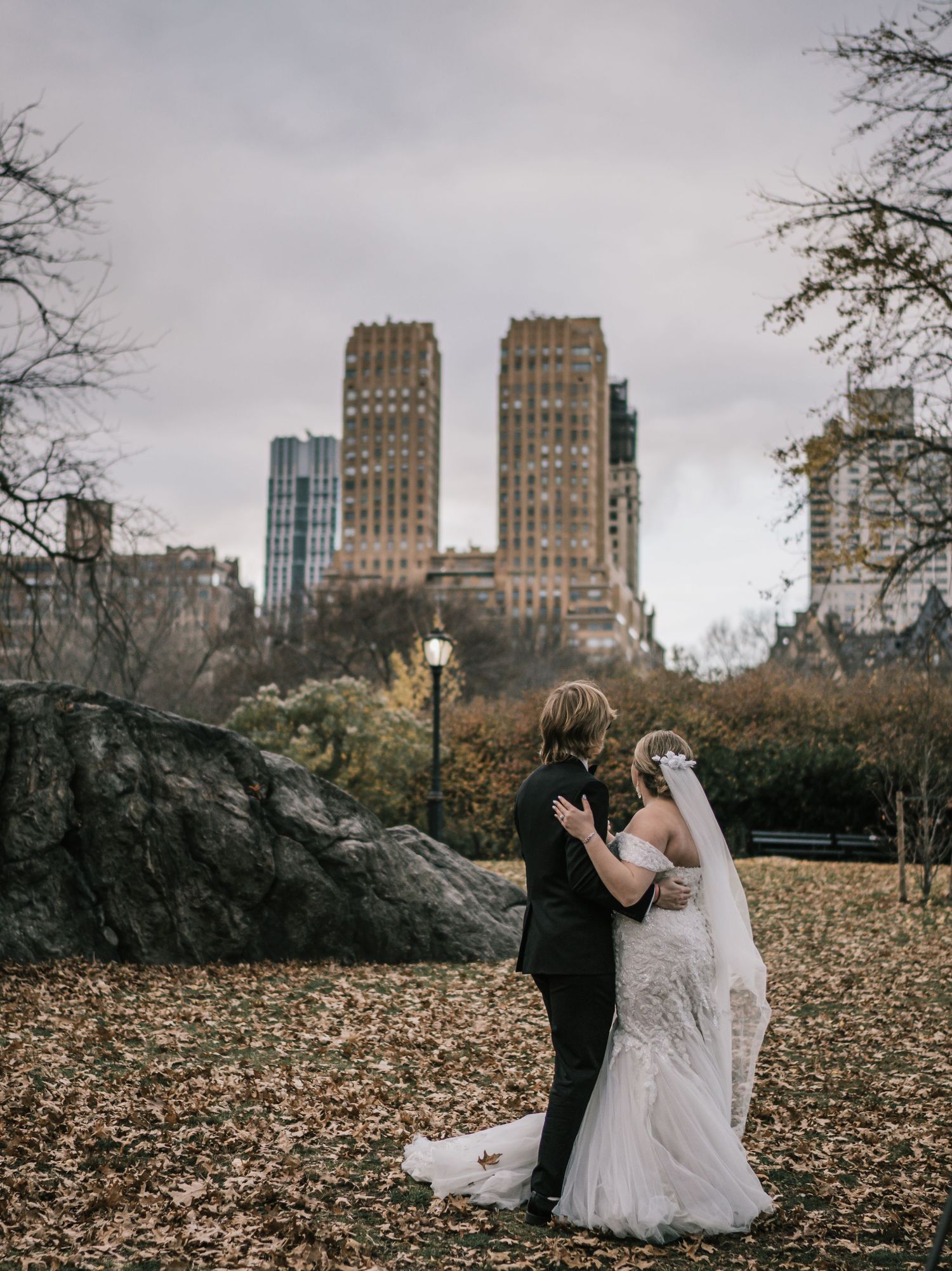 Bride and groom embrace in Central Park with NYC skyline and autumn foliage in background.