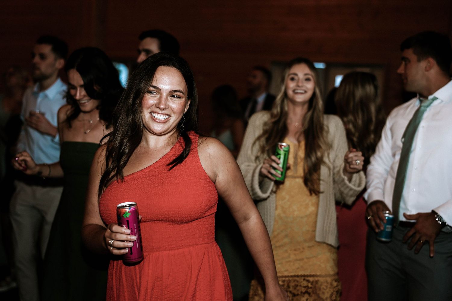 A smiling woman in a red dress holds a drink can while dancing at a wedding reception.