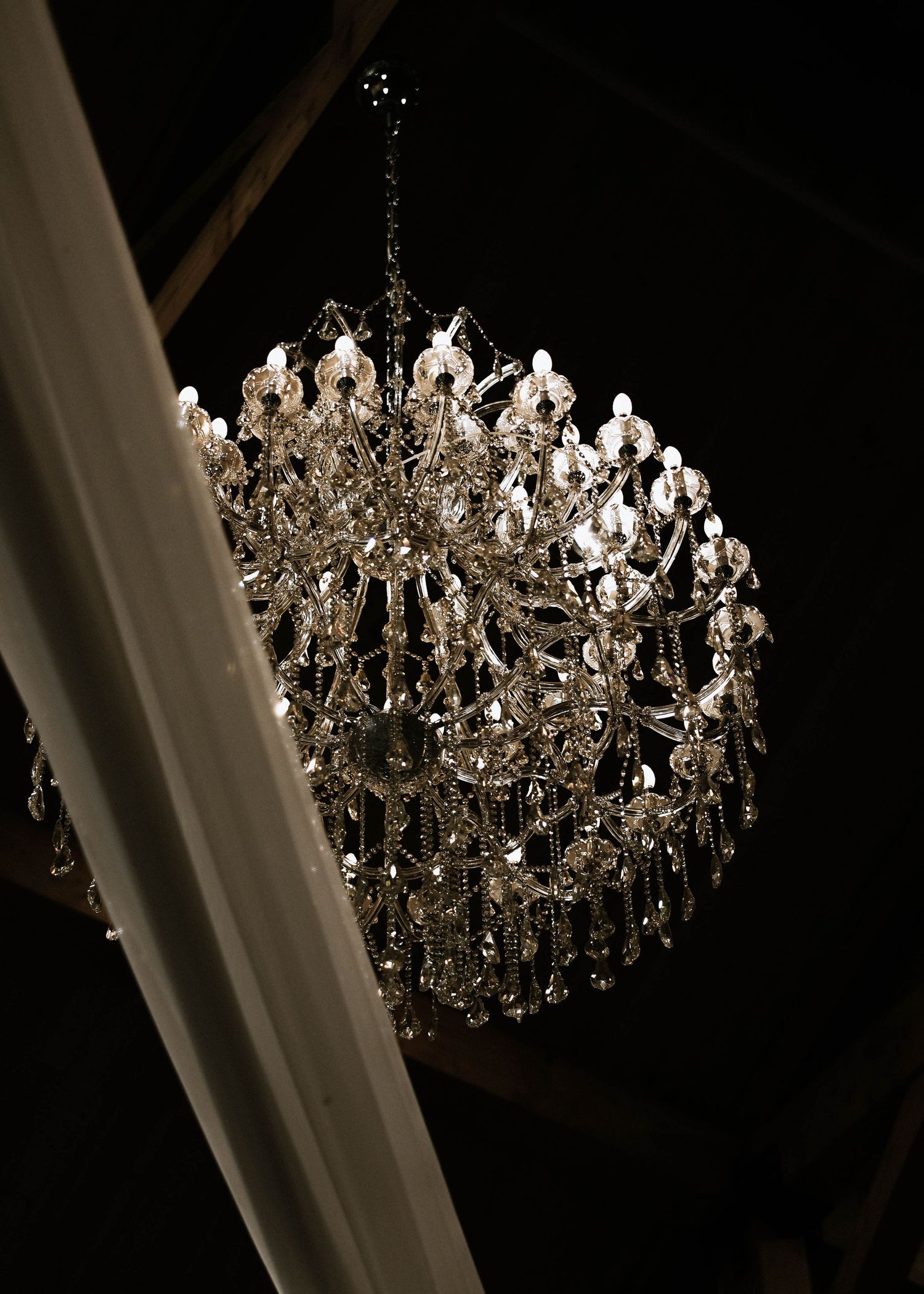 Elegant crystal chandelier with candle-style lights hanging from a dark ceiling at a wedding venue.