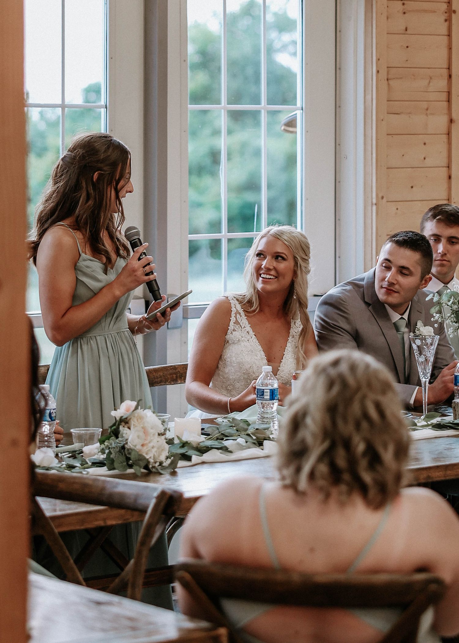 Maid of honor reads wedding speech into microphone as smiling bride listens at rustic barn reception.