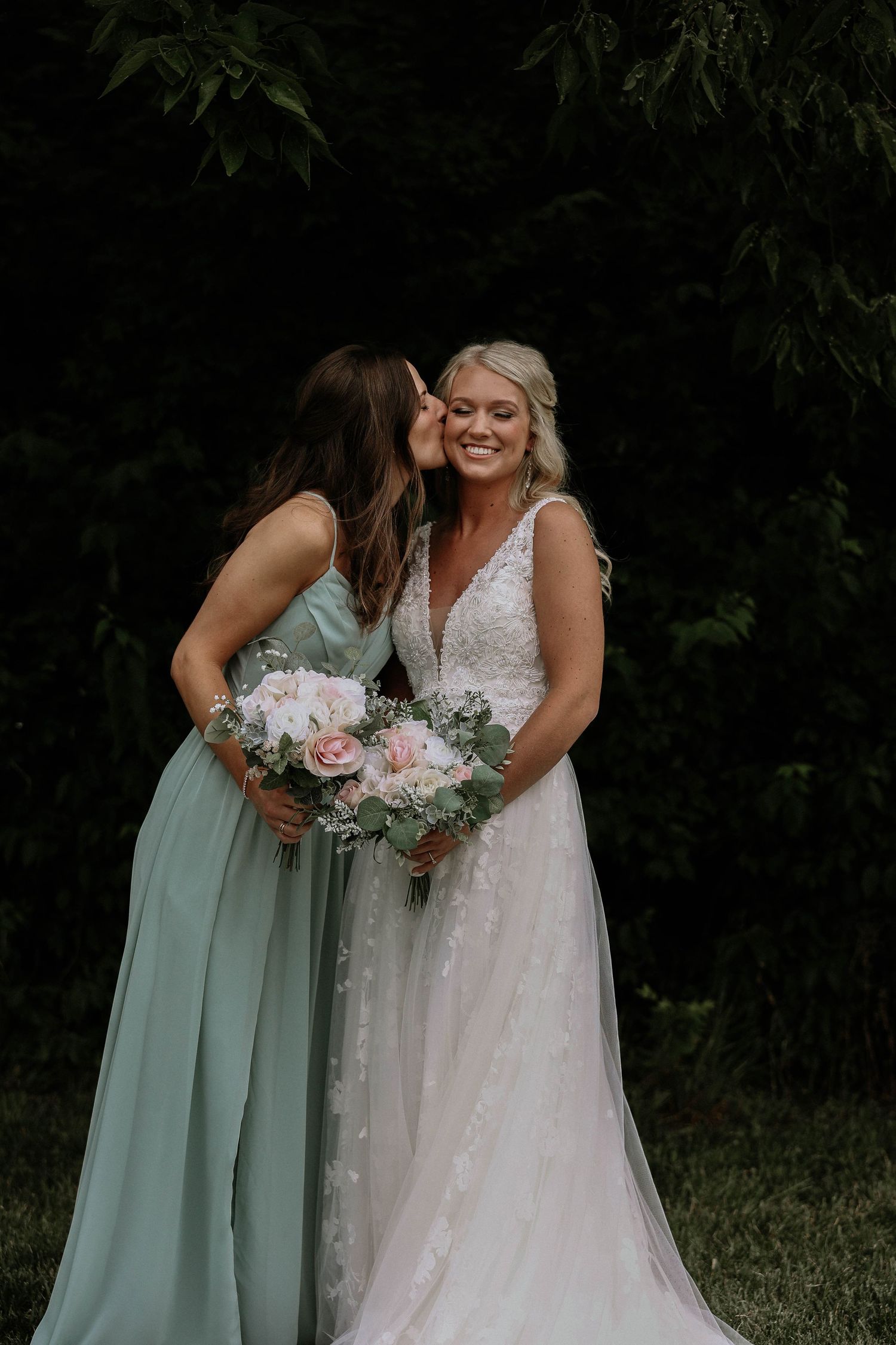 Bridesmaid in sage green dress kissing smiling bride in lace wedding gown while holding pink rose bouquet.