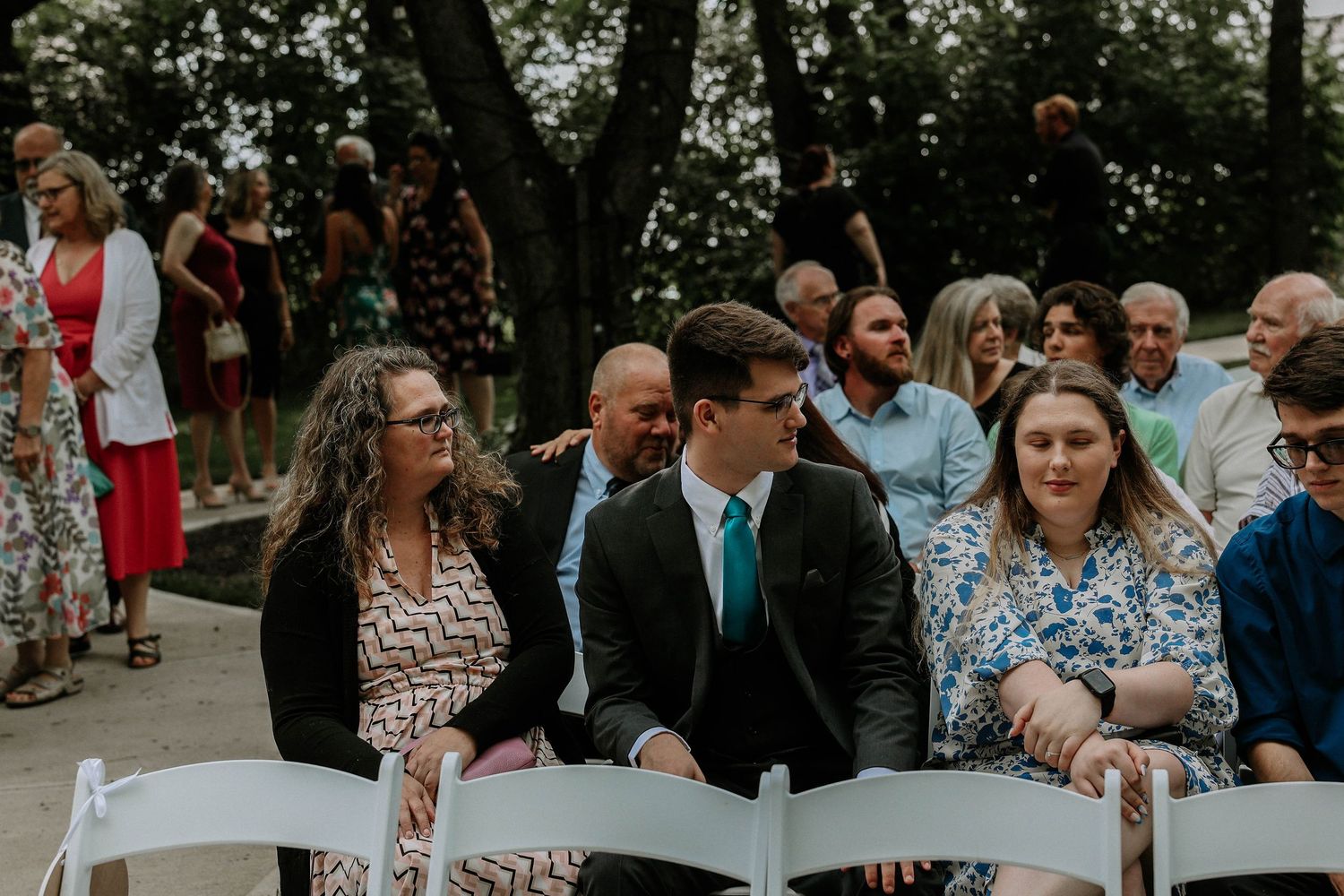 Wedding guests seated outdoors on white chairs, smiling and enjoying a ceremony in a garden setting.