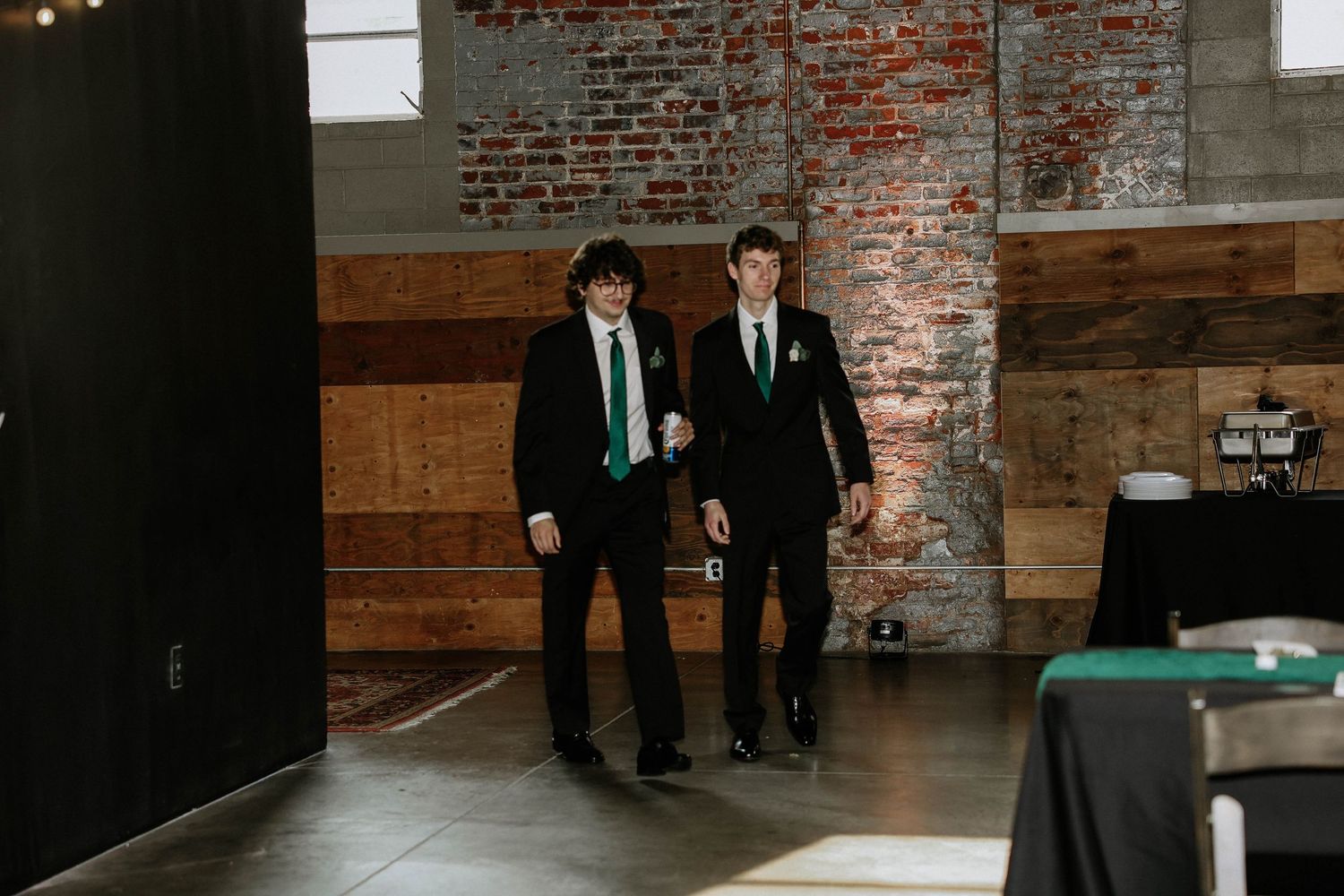 Two young men in black suits with green ties walking through an industrial venue with exposed brick walls.