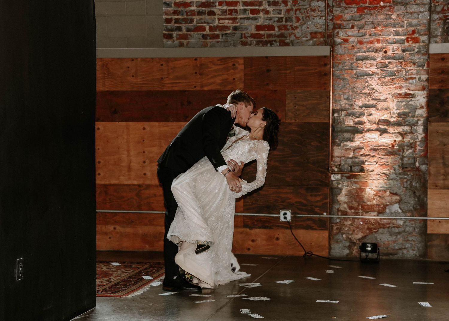Bride and groom share a romantic dip kiss on the dance floor at their wedding reception in a rustic brick venue.