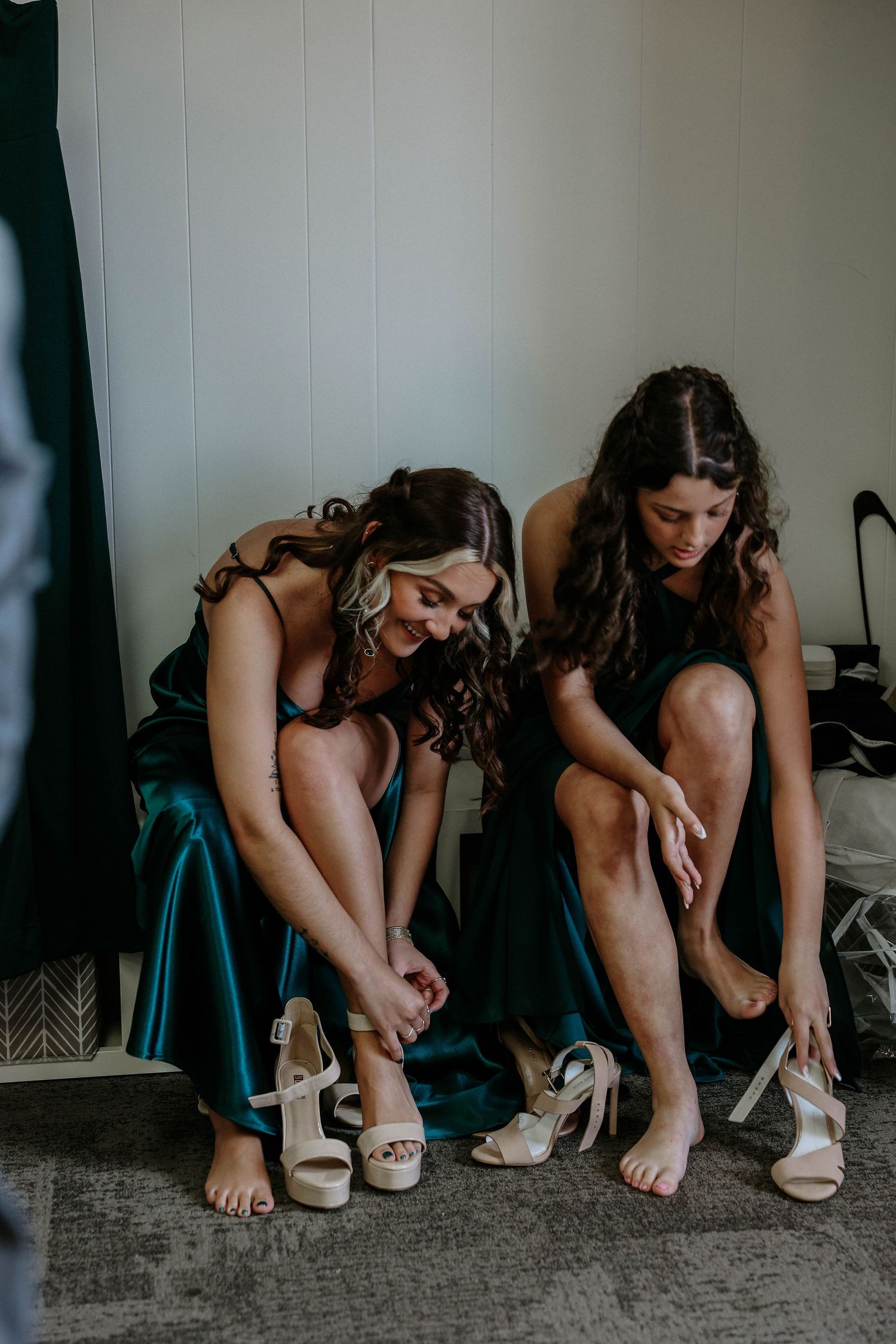 Two bridesmaids in green satin dresses sitting and putting on nude heeled sandals.