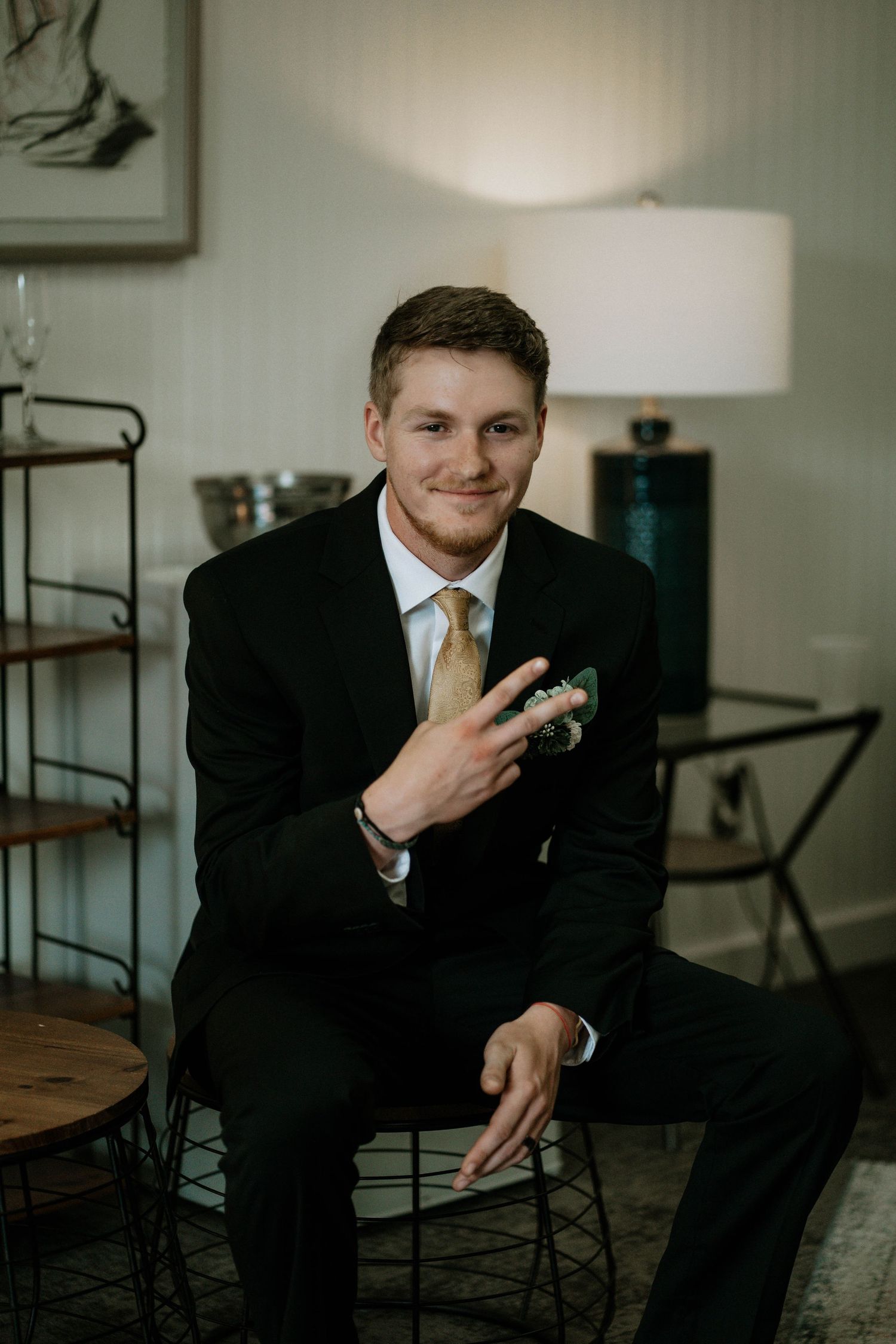 Dapper groom in black suit with gold tie sits smiling while adjusting his boutonniere before wedding ceremony.