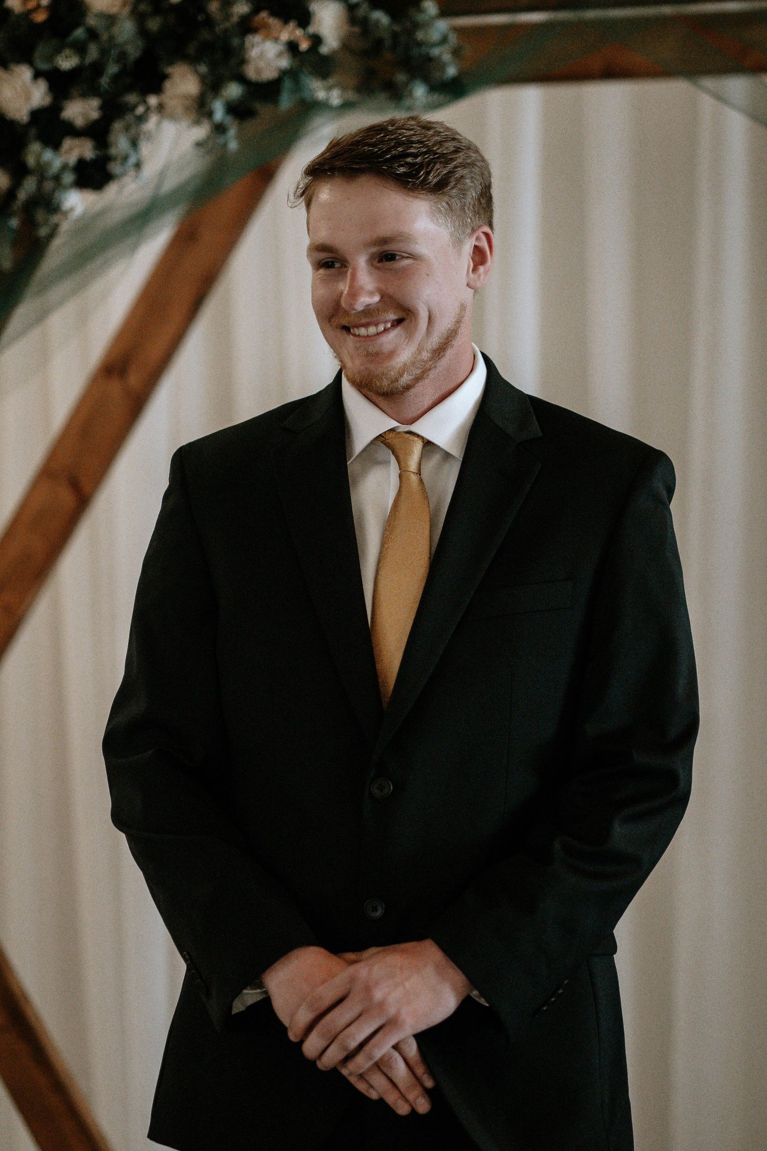 Smiling groom in black suit and gold tie standing at wedding altar with floral arch backdrop.