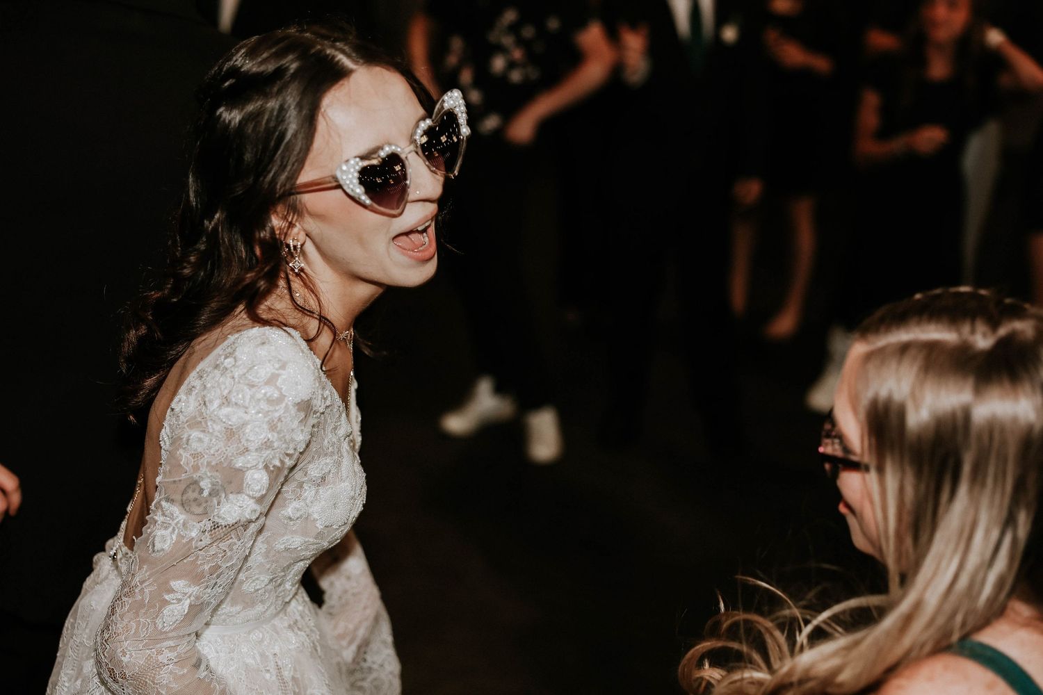 Bride in lace dress and heart-shaped sunglasses dancing with a guest at a wedding reception.