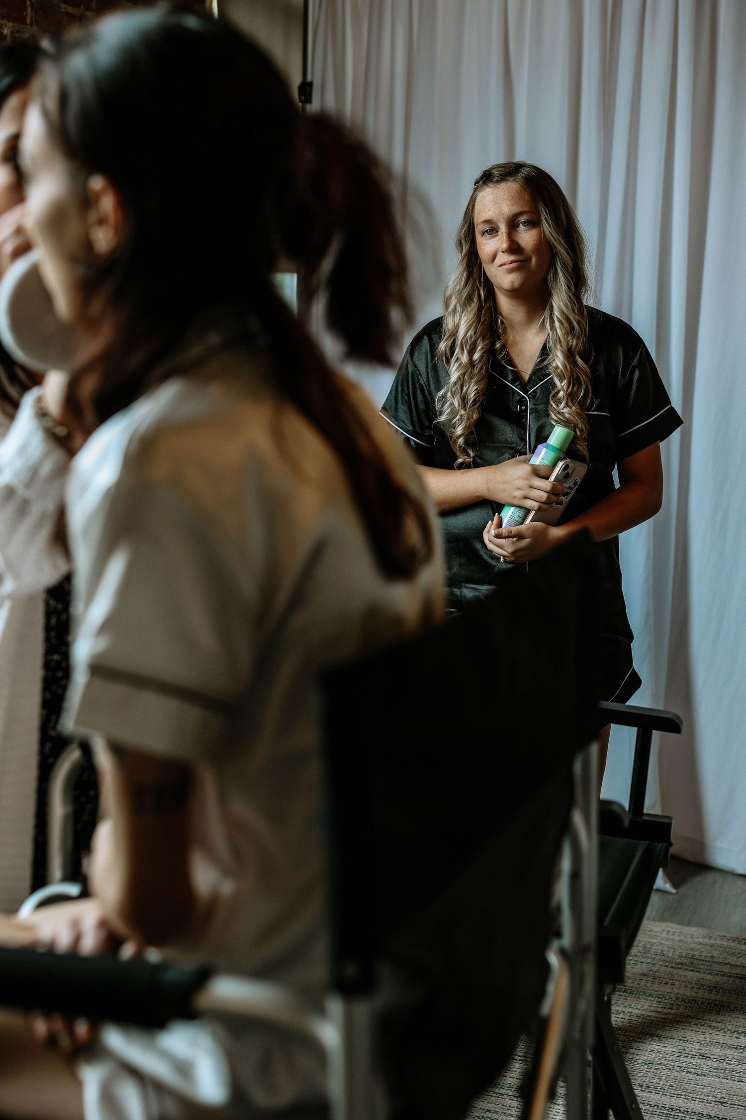 A smiling bridesmaid in black satin pajamas holds hair products during bridal preparations.