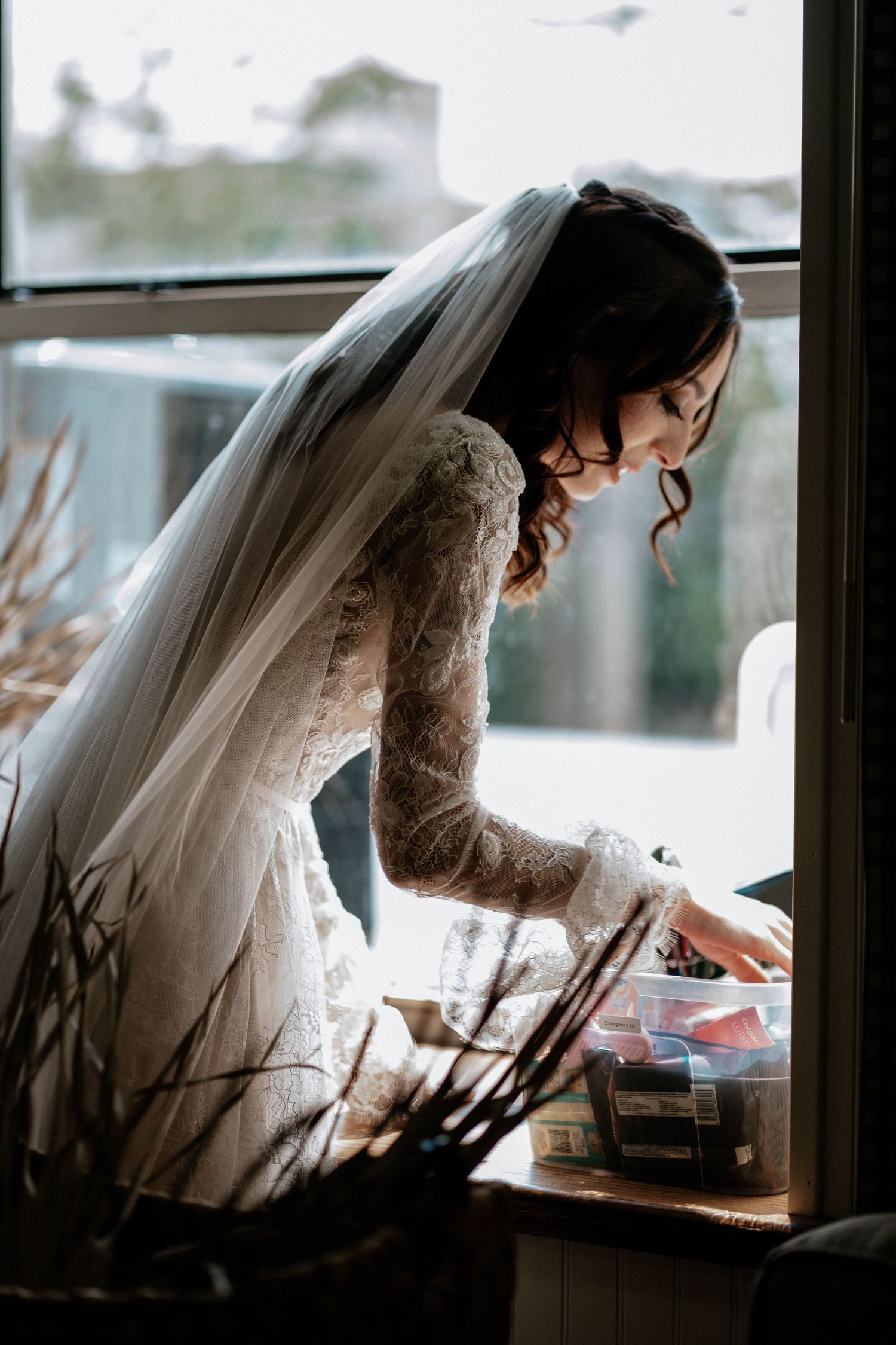 Bride in lace long-sleeve gown and veil leans over a table by a window on her wedding day.