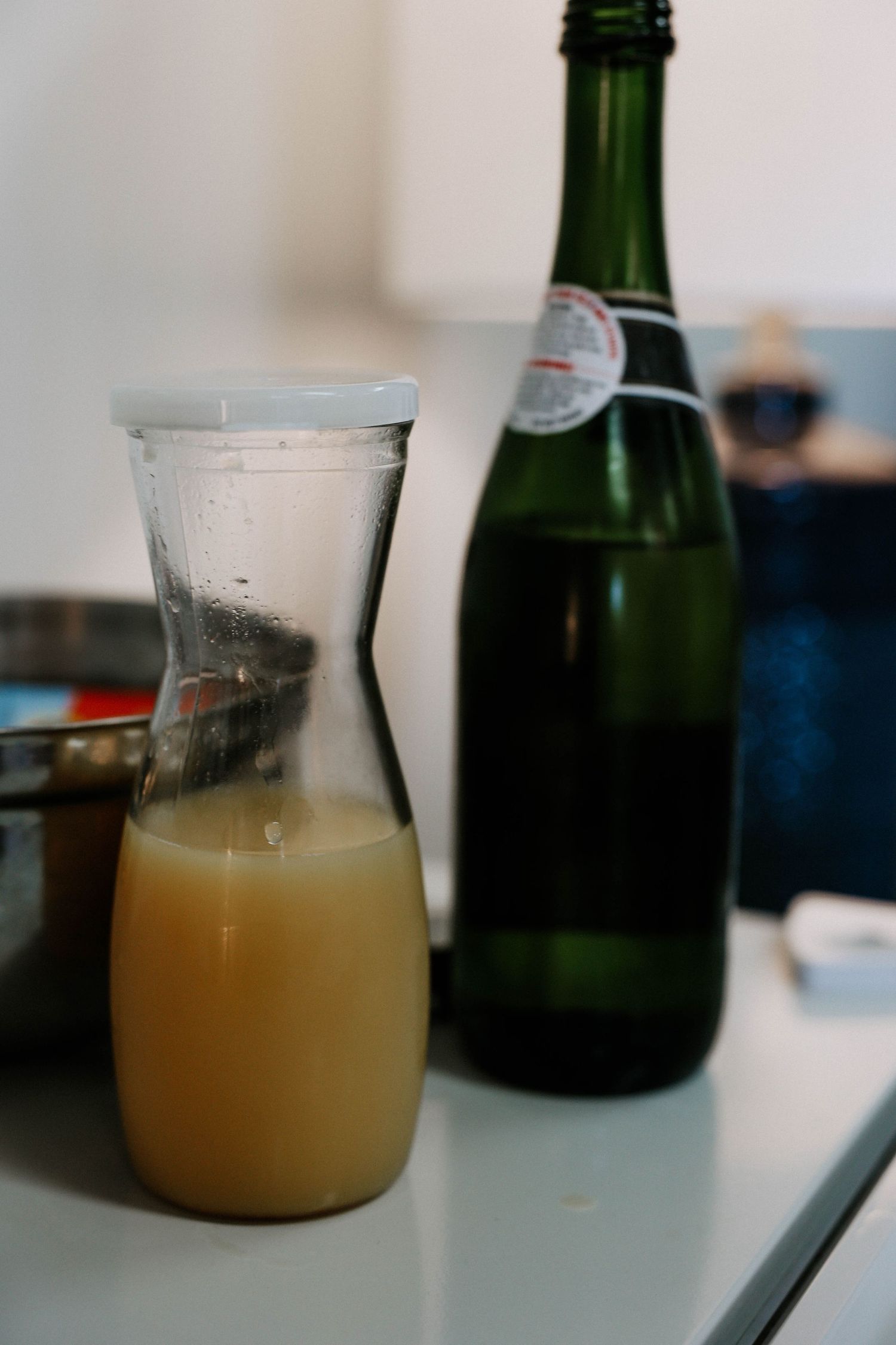 A glass carafe of fresh juice beside a green wine bottle on a light blue table.