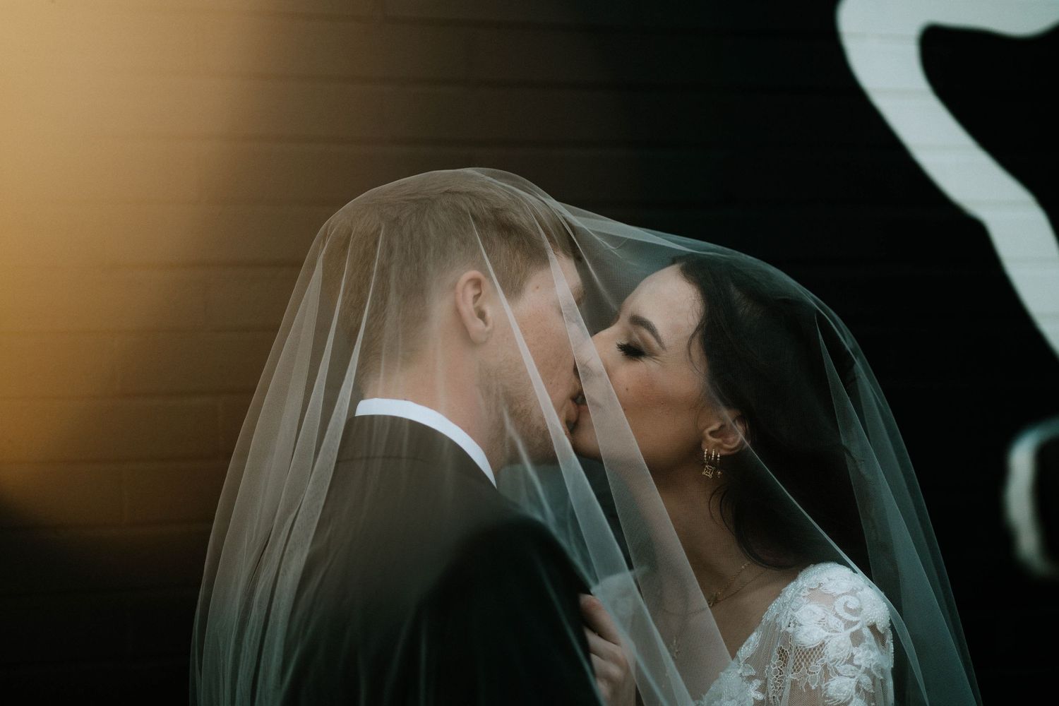Bride and groom sharing a romantic kiss under a flowing white veil on their wedding day.