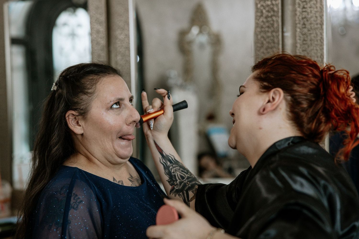 A makeup artist with red hair and tattoos applies makeup to a woman in a blue dress at an elegant venue.
