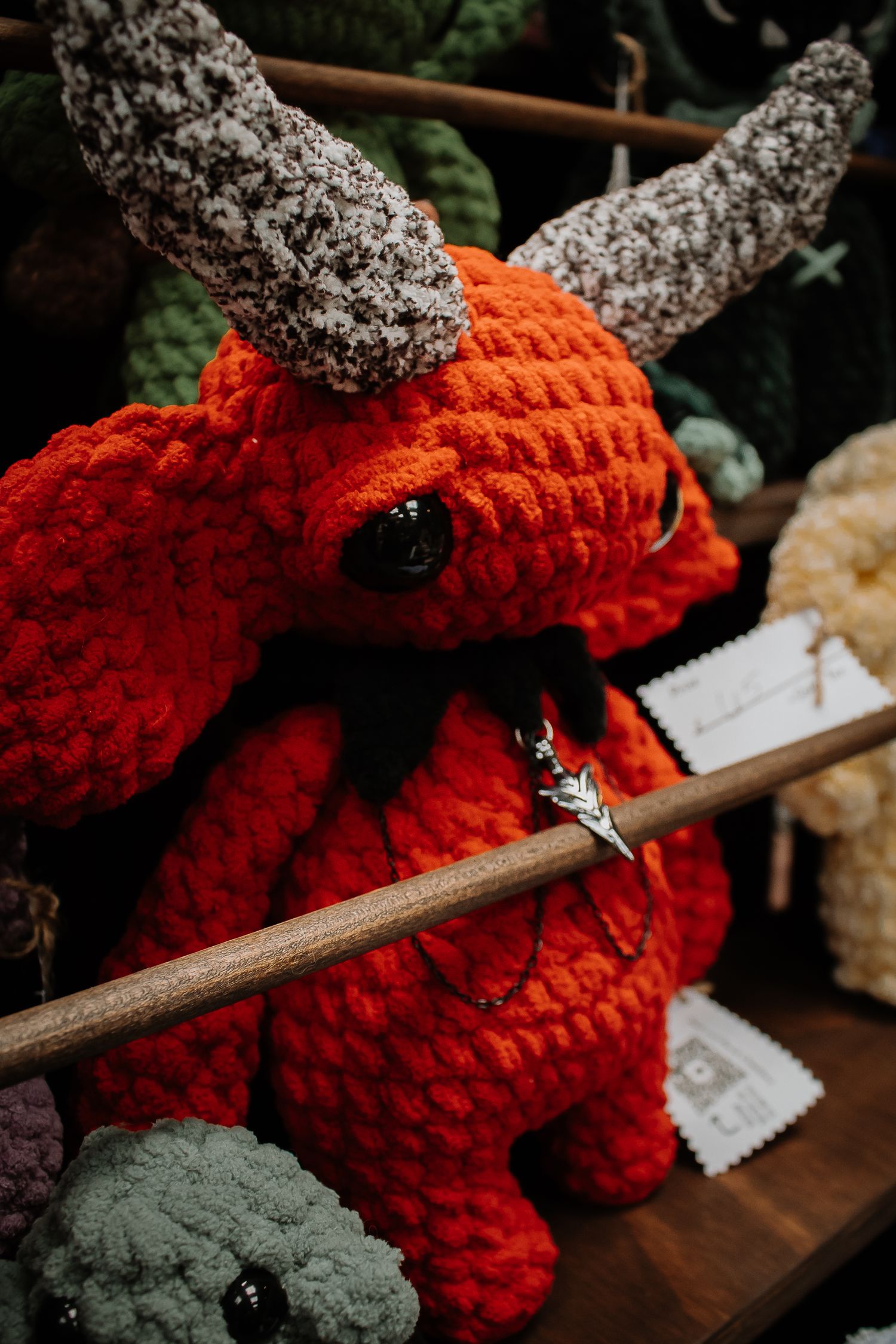 Red crocheted devil amigurumi with horns and pentagram necklace displayed on wooden shelf at craft market.