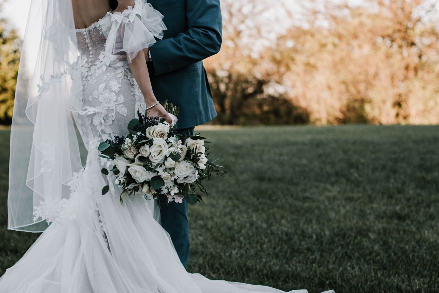 Bride in lace gown holds white floral bouquet while standing with groom on green lawn at sunset at Stone Valley Meadows in Dayton, Ohio