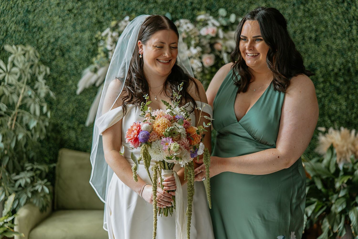 Smiling bride holding a colorful wildflower bouquet stands with her bridesmaid in sage green dress.