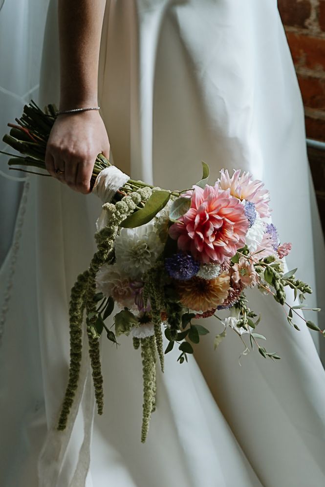 Bride holding a lush cascading wedding bouquet with pink dahlias and wildflowers against white gown.