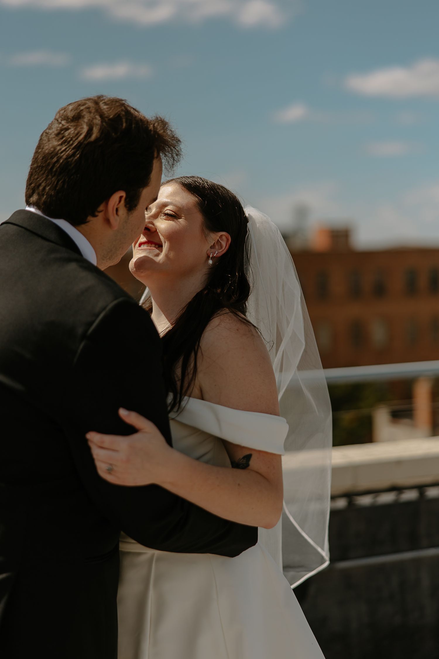 Joyful bride laughs while embracing groom on sunny rooftop with brick building in the background.