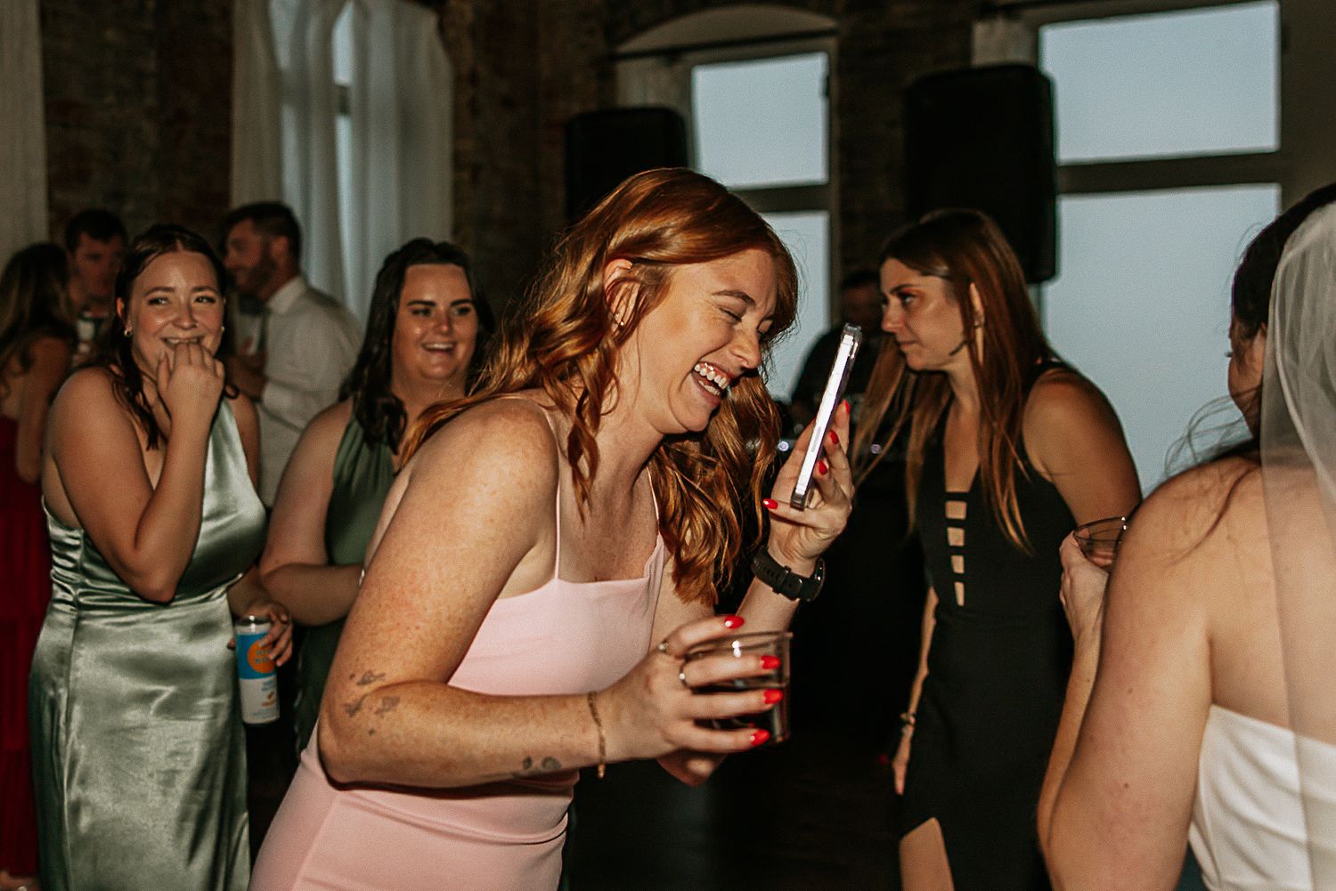 Guests laugh and socialize at a wedding reception, with a redhead holding a red cup.