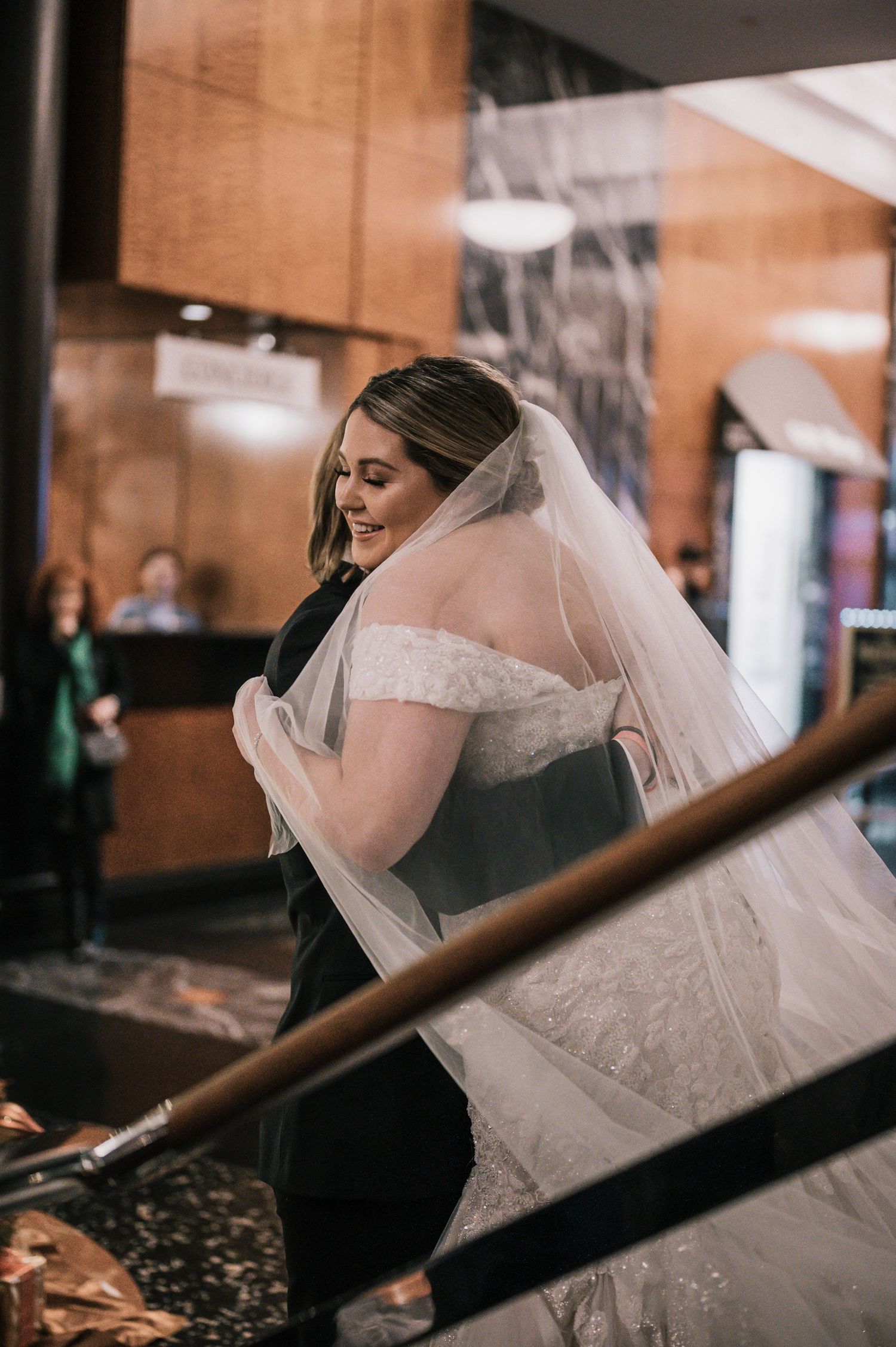 Bride in lace mermaid gown with flowing veil embracing groom at indoor wedding venue.