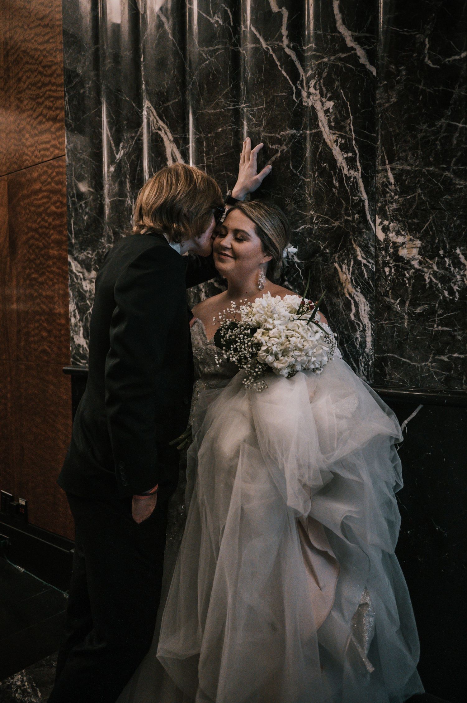 Bride and groom in intimate embrace against dark marble wall, bride holding white floral bouquet in NYC.