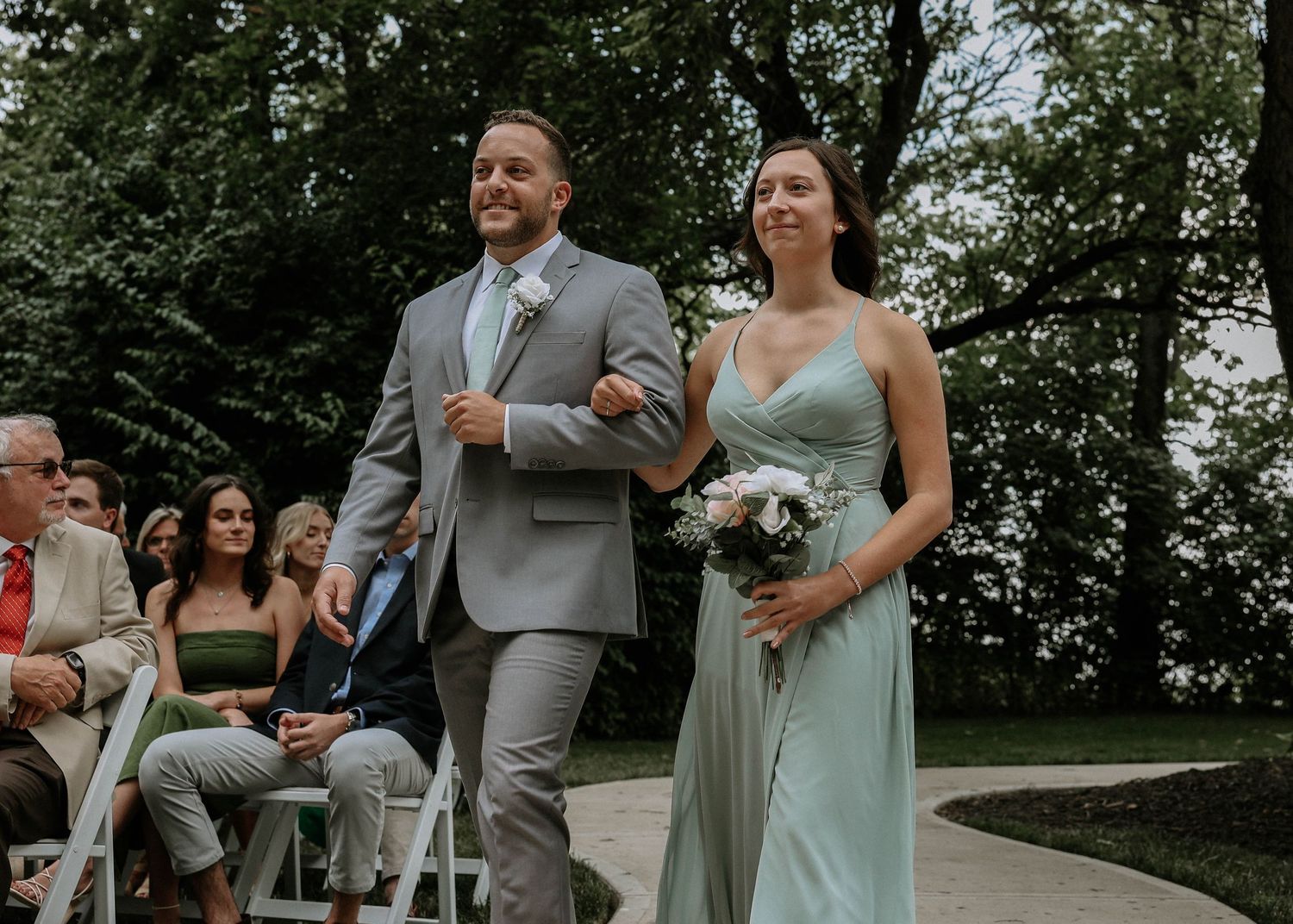 A groomsman in a gray suit escorts a bridesmaid in a sage green gown down an outdoor wedding aisle.