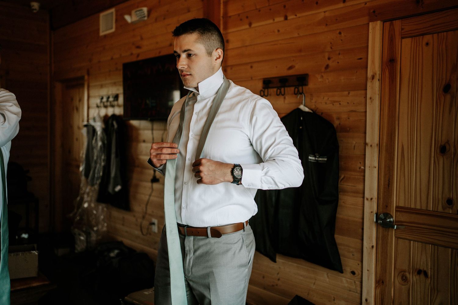 Groom getting dressed in a wood-paneled room, adjusting his tie before the wedding ceremony.
