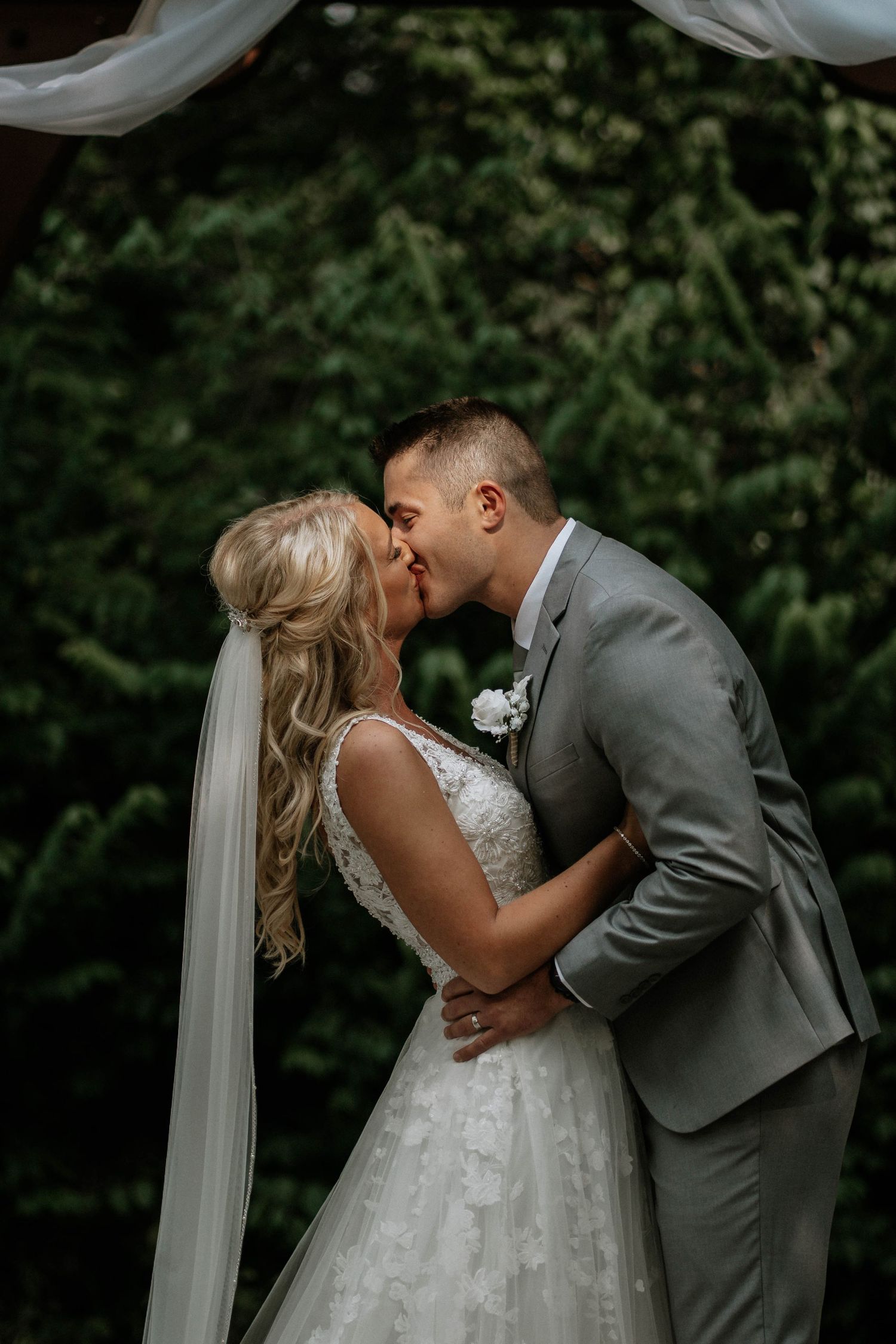 Newlyweds kissing under floral arch surrounded by lush greenery, bride in lace dress with long veil.