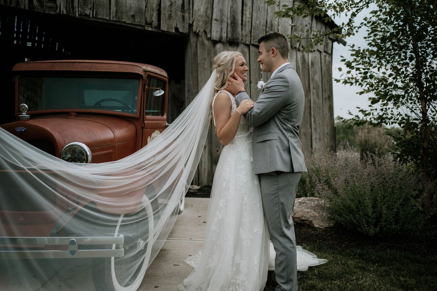 Bride and groom sharing intimate moment near vintage red truck and old barn, veil flowing.