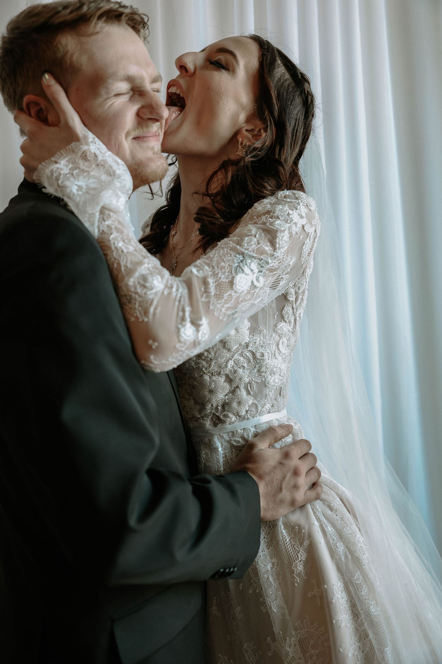 Joyful bride in lace gown laughing while embracing groom in dark suit near white curtains.