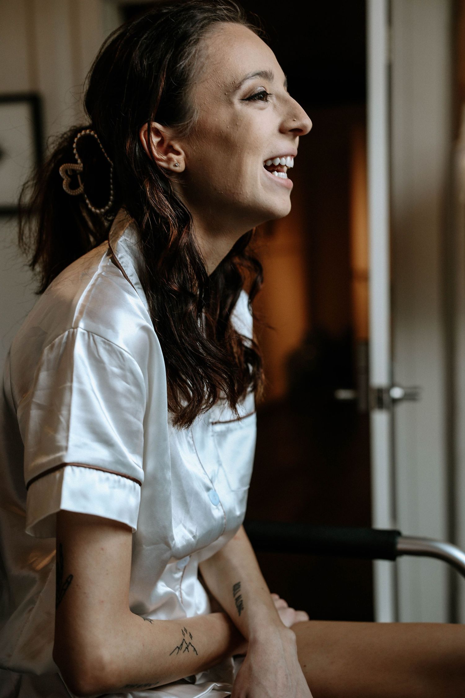 A laughing bride in white satin pajamas with a tattoo on her arm getting ready on her wedding day.